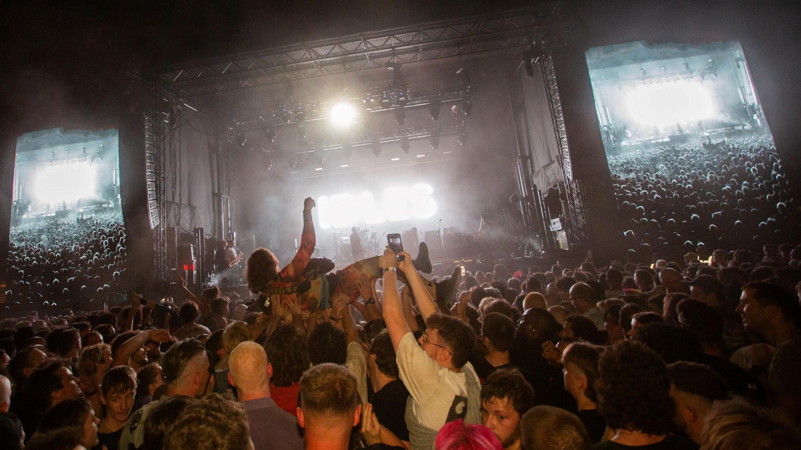 A member of the band Idles is held aloft by the crowd in Queen Square in Bristol as he plays a guitar. In the background the stage is visible, along with two large screens either side which are showing the crowd