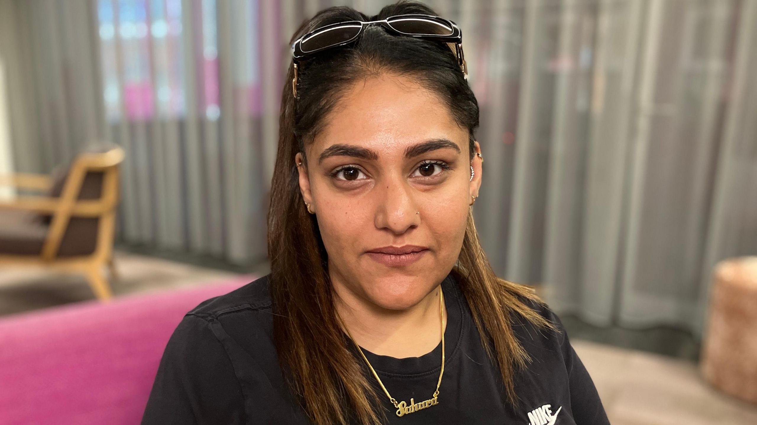 A woman looks directly into the camera - she has big brown eyes, a ponytail with long dark hair, pair of glasses on her head, a black t-shirt and gold necklace with the word 'Sahara', the name of her daughter who died.
