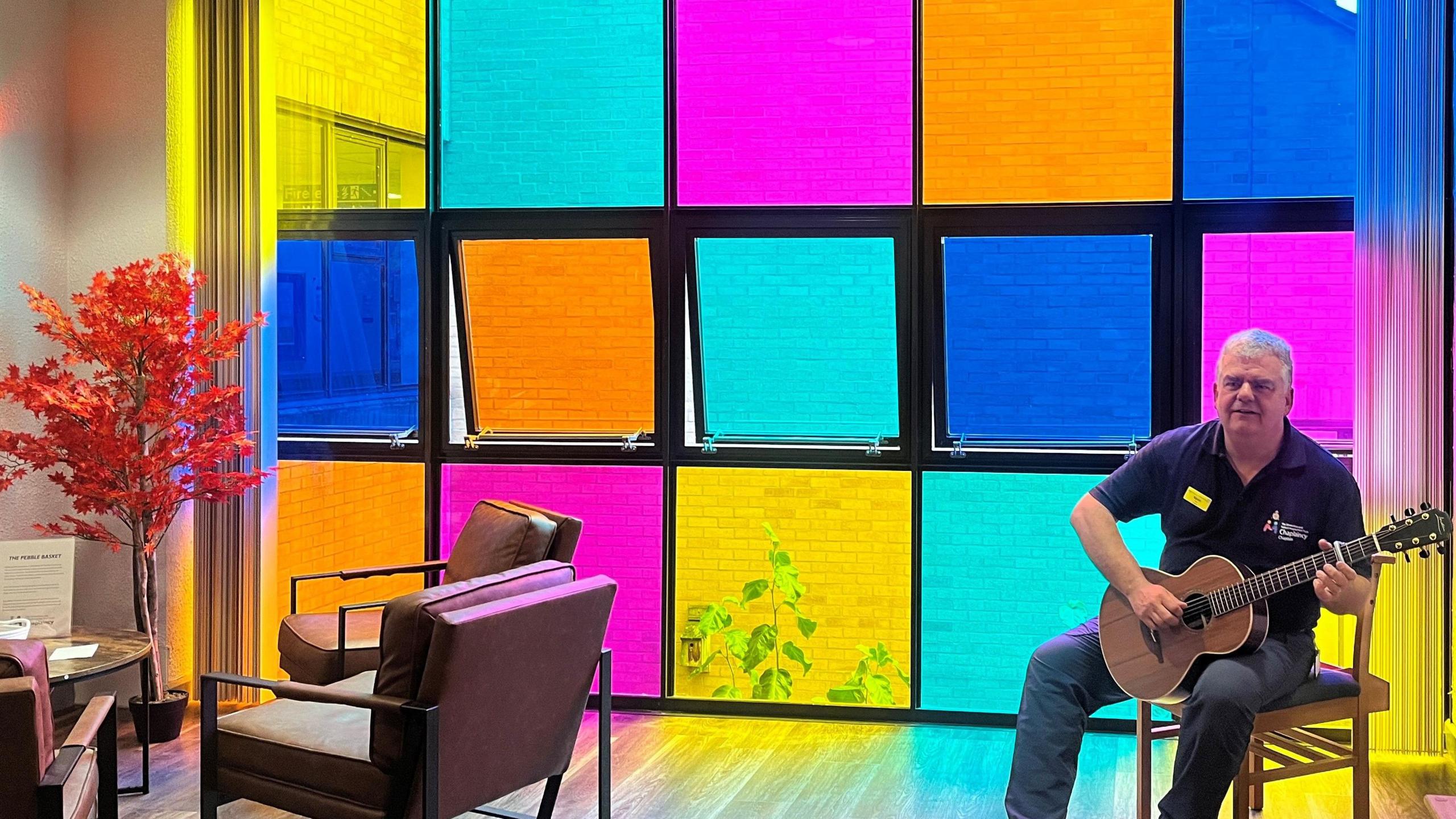 Father Kevin Place, wearing blue jeans and tshirt, is sitting on a chair playing the guitar. Behind him is a large window made up of 15 squares of brightly coloured glass, including blue, yellow, pink and orange. To Kevin's left is three brown chairs, a plant and a small table.