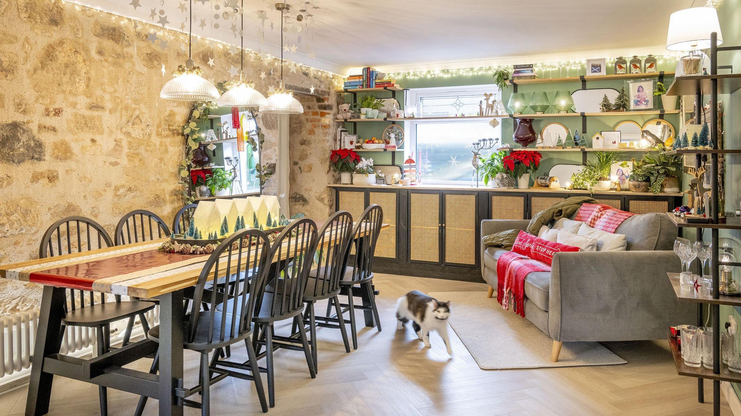 A brightly lit festive kitchen. Star ornaments dangle above a long dining table which is decorated with a model Christmas village. The walls are covered in shelves which hold a mix of ornaments and crockery. A brown and white cat wanders across the floor.