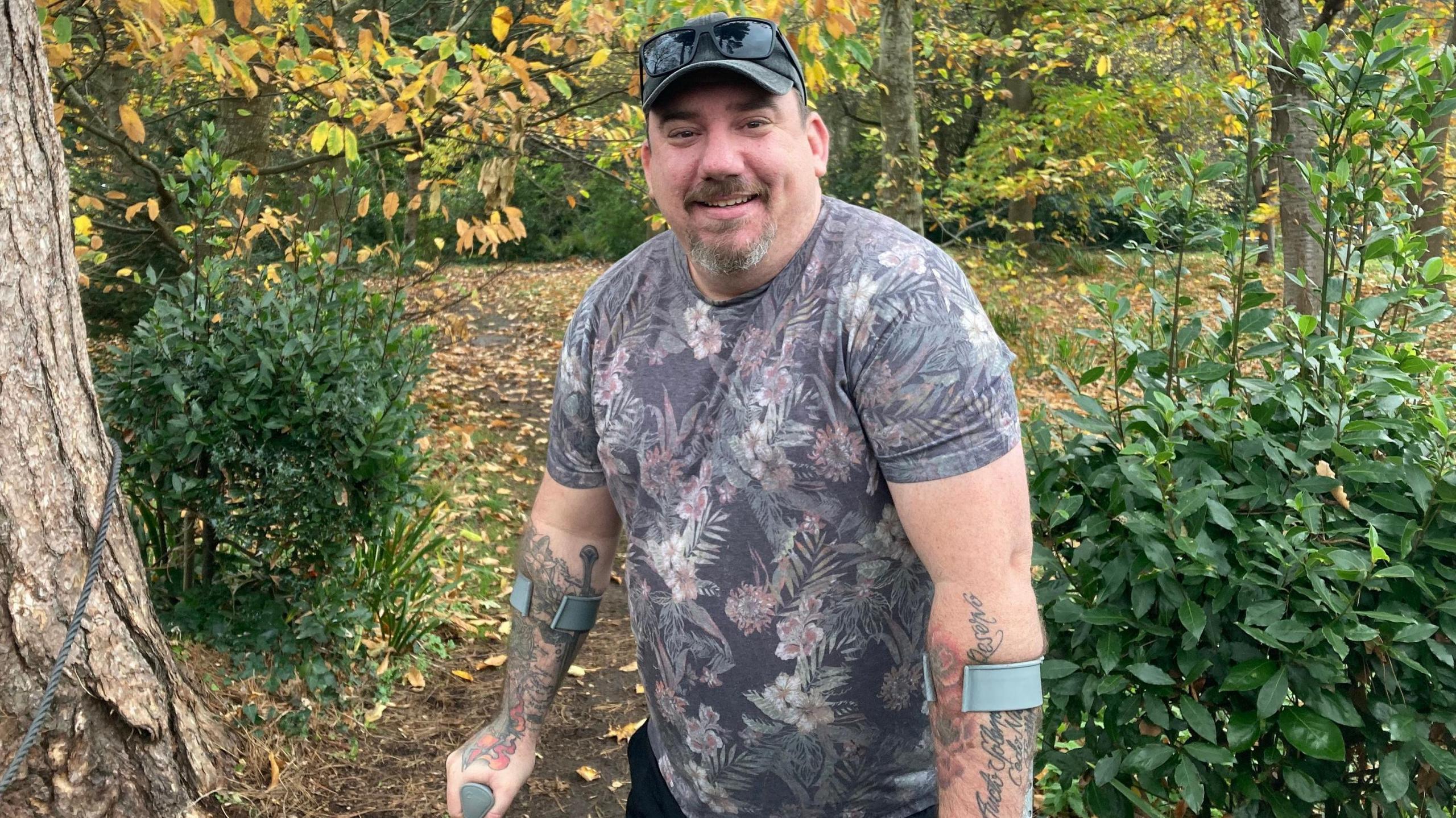Dan Stone standing in Woodland smiling at the camera. He is wearing a flowery T-shirt and has tattoos on his forearms. He also has a cap and a short goatee beard. He is using crutches because of his knee injury.