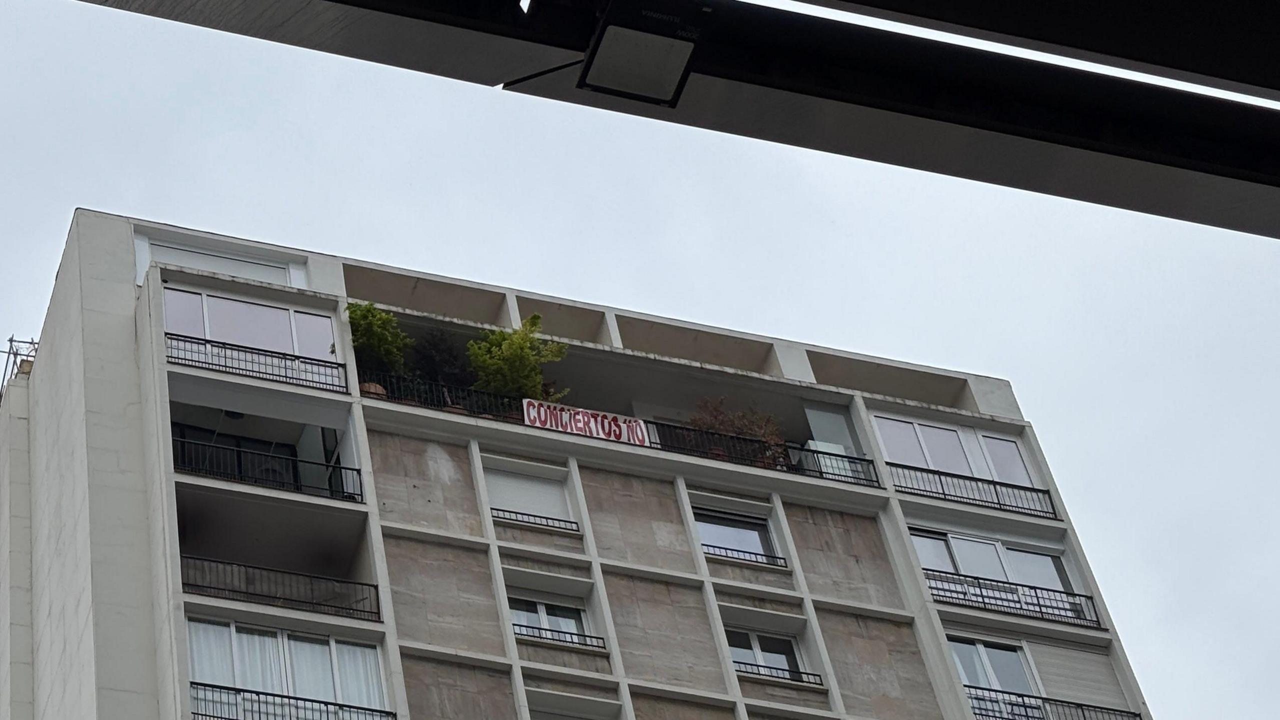 A high rise flat building. Grey façade with a balcony full of plants. from the railing hangs a banner reading no concerts in Spanish. It is written in big red letters on a white background.