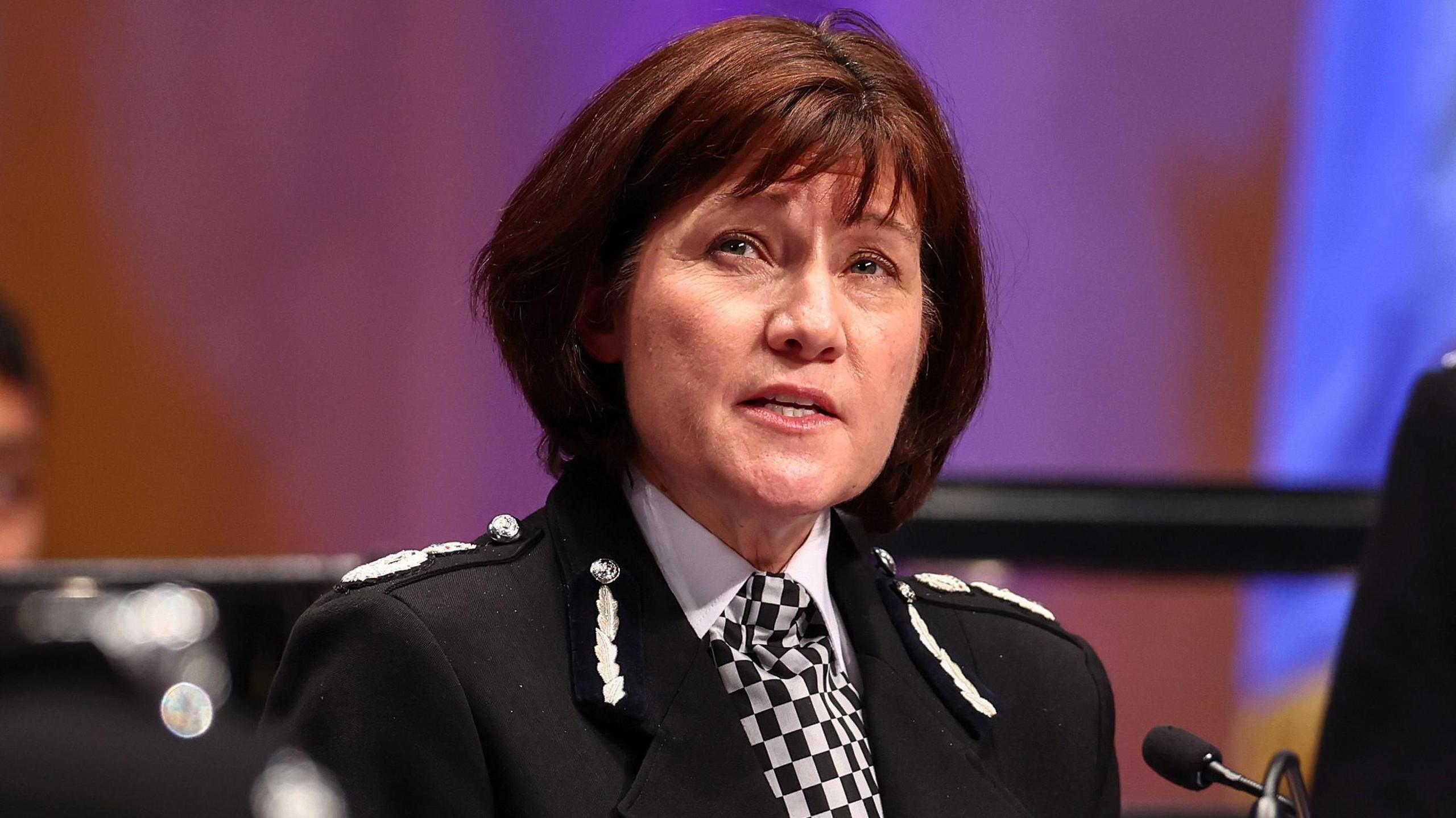 Head and shoulders shot of chief constable Jo Farrell. She is wearing a black police jacker with white braids, a white blouse and a black and white checked cravat. She is looking off into the distance with a serious expression on her face. A maroon and blue backdrop can be seen in the background.