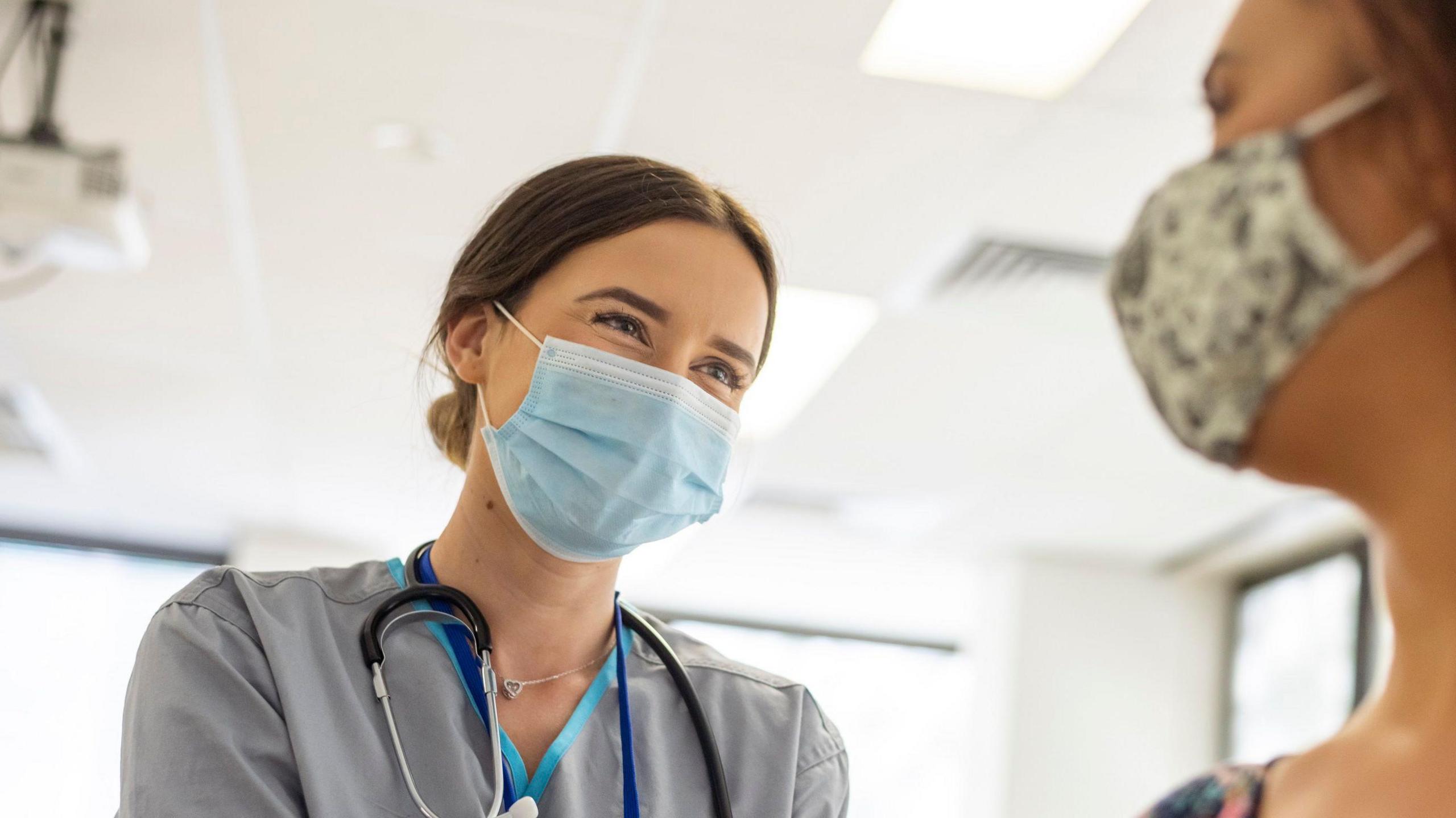 Doctor wearing blue face mask while looking at patient with flowery mask on. They are sat inside a white hospital room.