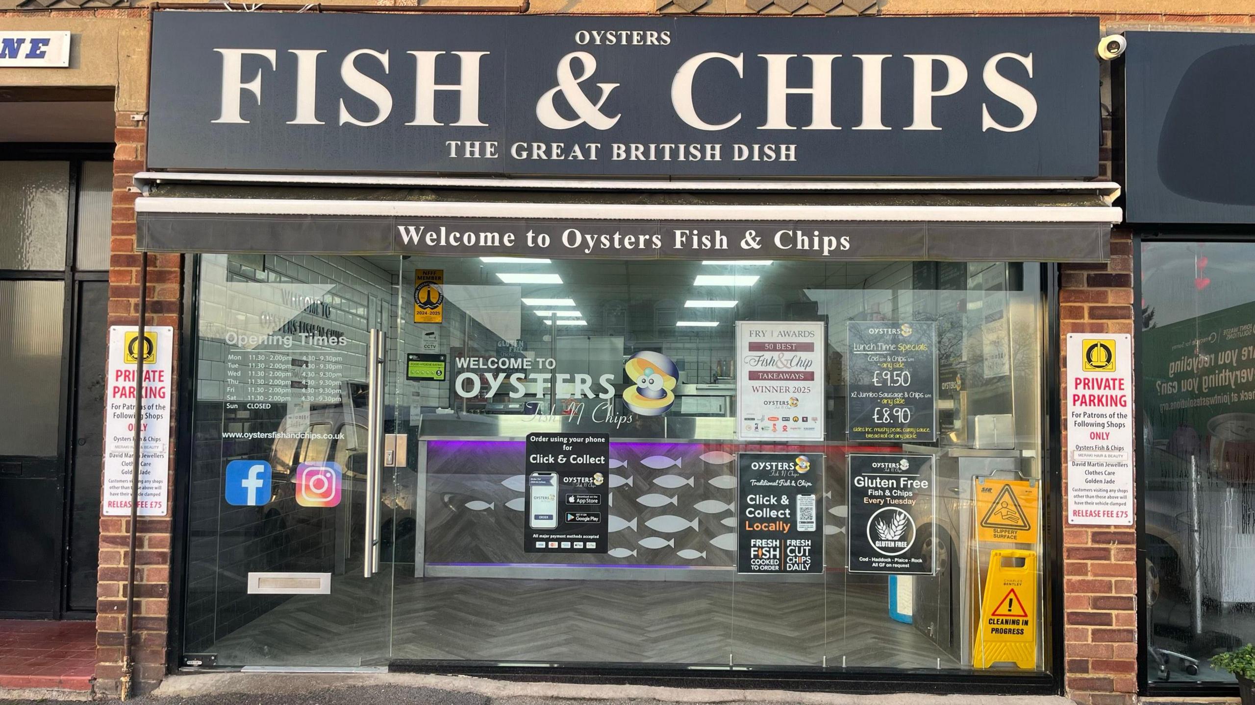 A fish and chip shop with a black sign above it and a glass exterior. The shop sign reads "Oysters / Fish & Chips / The Great British Dish". The glass window of the shops has a number of small signs reading "welcome to oysters" as well as prices and social media logos.