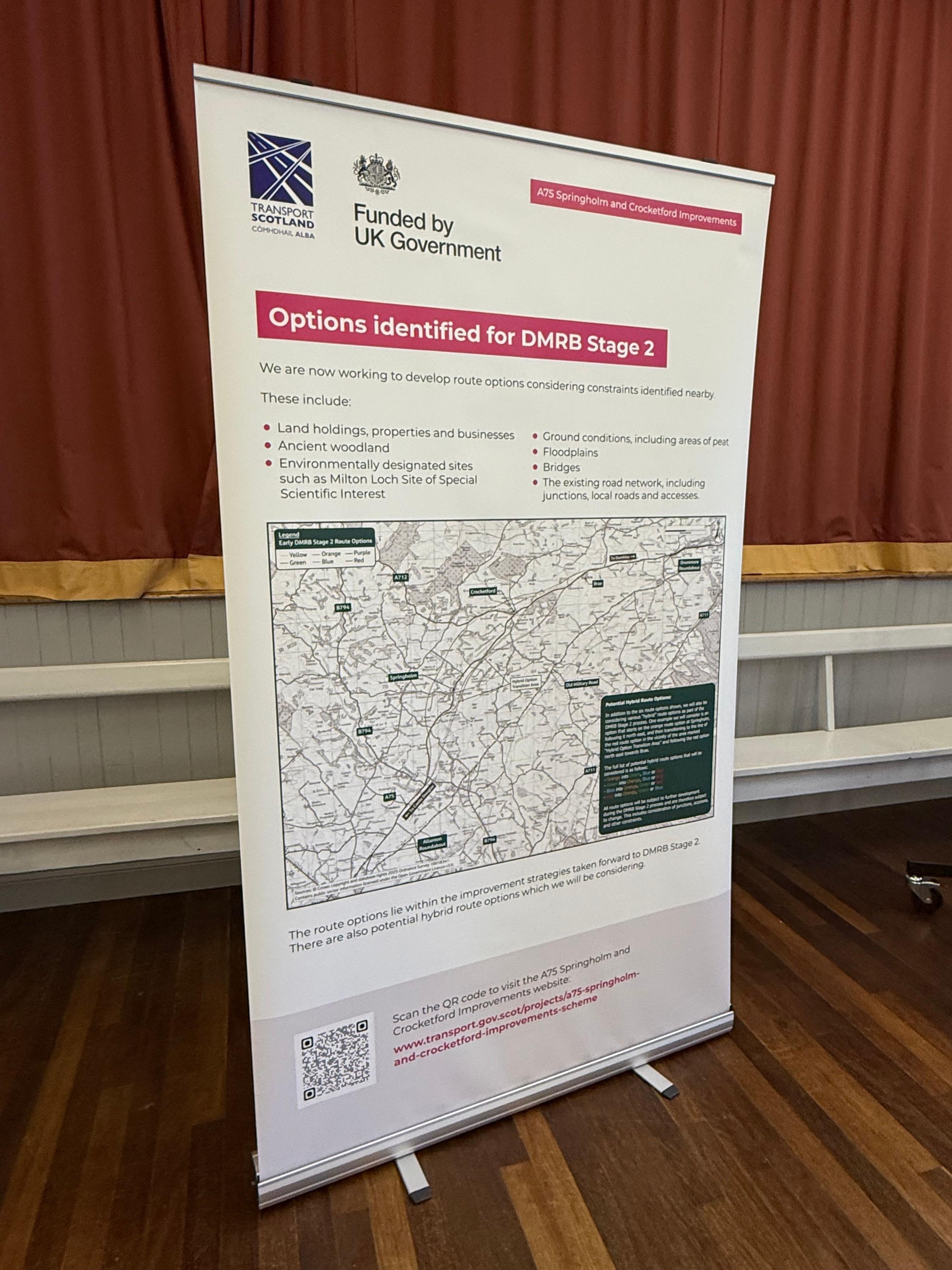 A banner type poster on a stand showing a map with potential options for a bypass on the A75 in the south of Scotland. It stands on a wooden floor with a large velvet-looking curtain behind it.