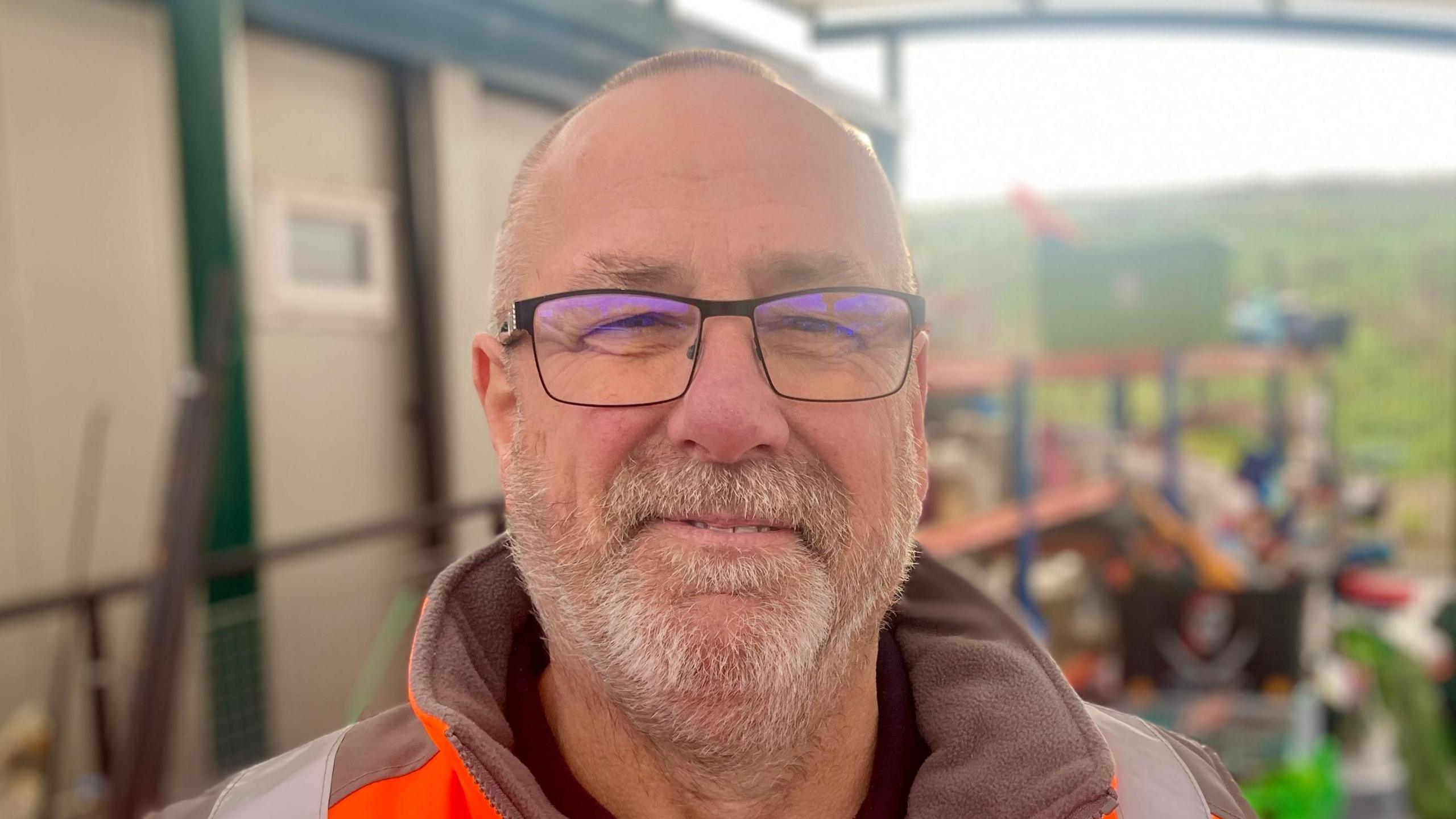 A man with thin light hair, wearing black glasses as well as an orange hi-vis coat, smiles at the camera. He has a grey and blonde beard. 