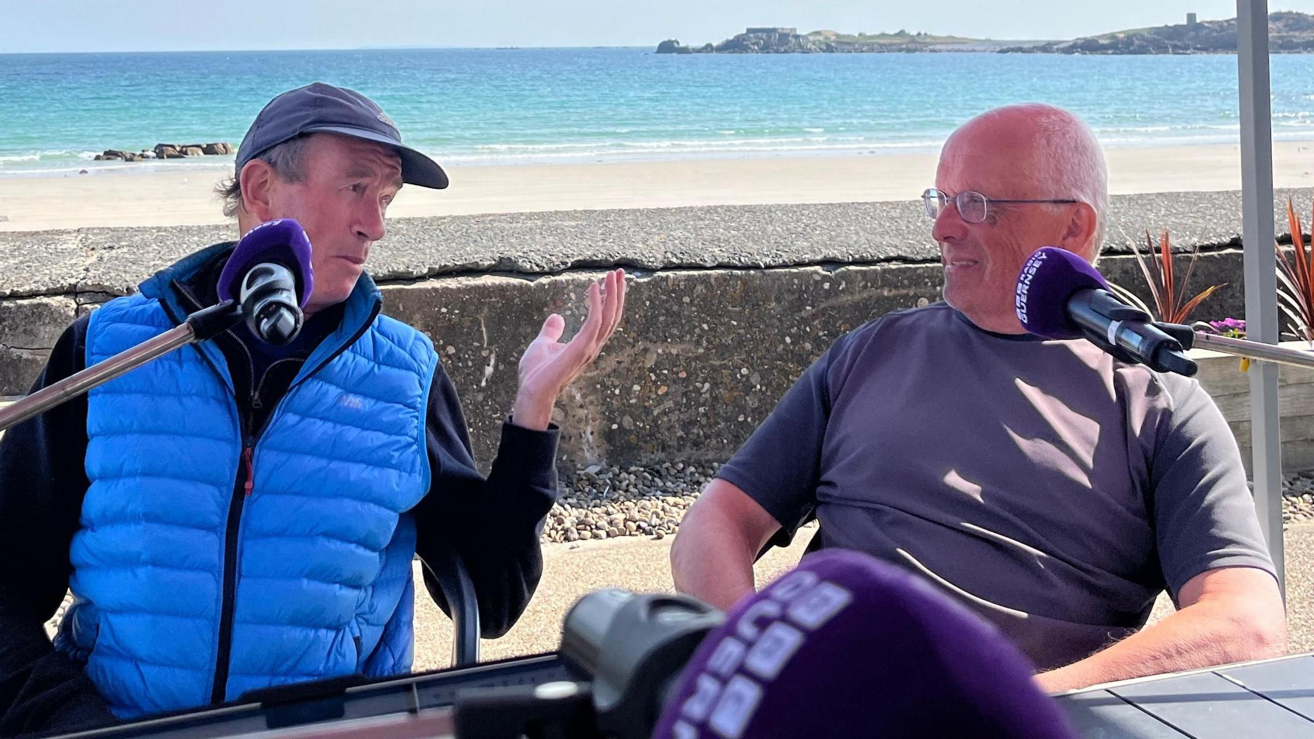 Two men in front of microphones discussing politics, the man on the left is wearing a blue gillet and is gesticulating towards the man on the right. They are in front of a beach backdrop on a sunny day. 