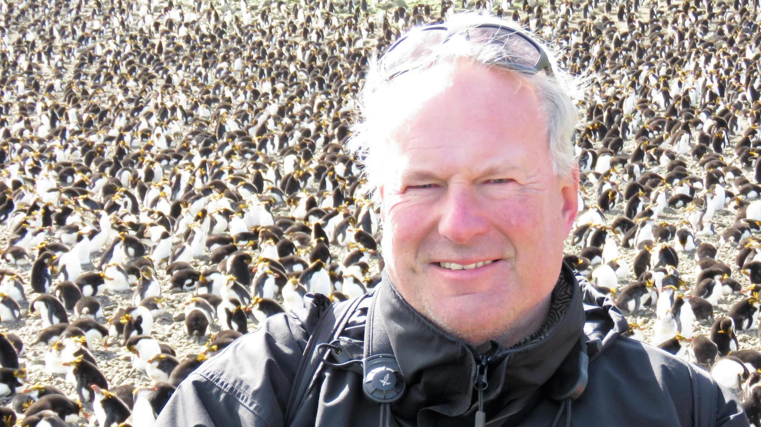 Keith Betton, who is wearing a black coat and has a camera around his neck, is smiling for a posed picture in front of colony of penguins. 