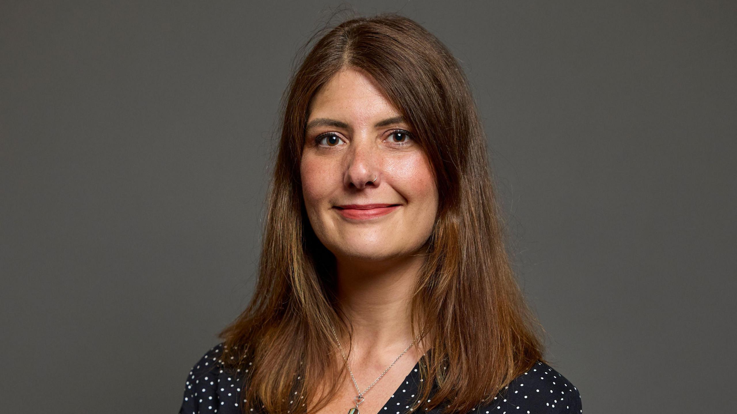 MP Cat Eccles, a woman with brown hair, is stood looking at the camera and smiling. She is wearing a necklace and a black top with white dots.