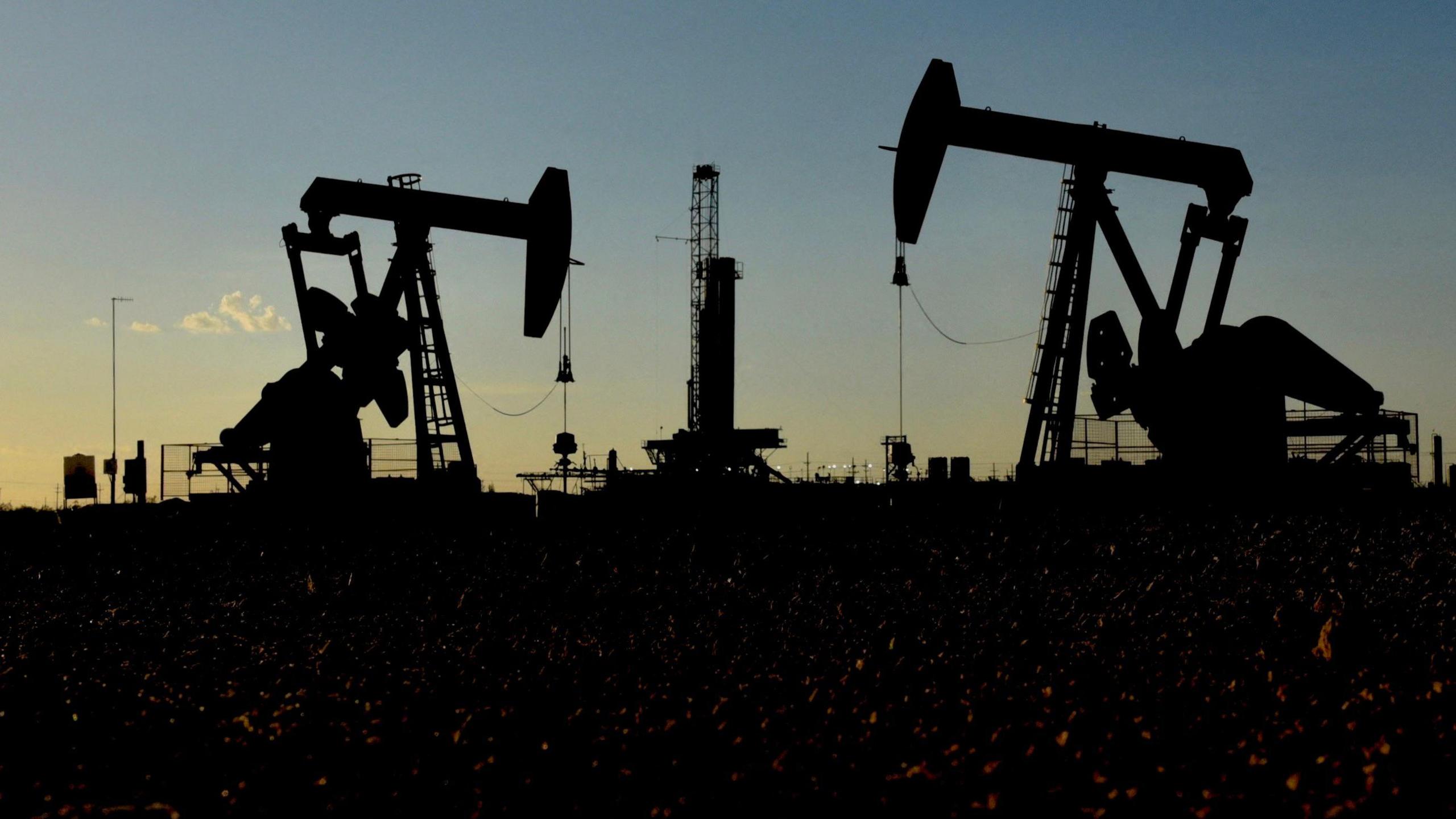 Image shows two pump jacks in a Texan oil field, silhouetted against the setting sun. 