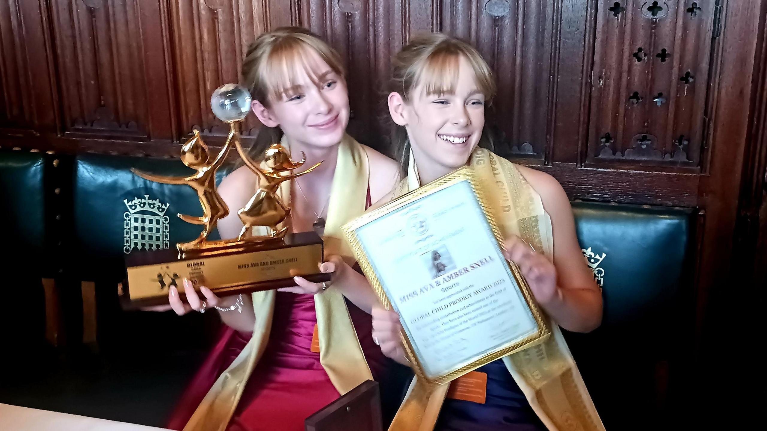 A pair of teenage girls who are identical set of twins, both blonde and wearing gold scarves over their necks, holding up a gold award and a framed certificate. They are sitting on green-cushioned chairs with the Houses of Parliament logo.