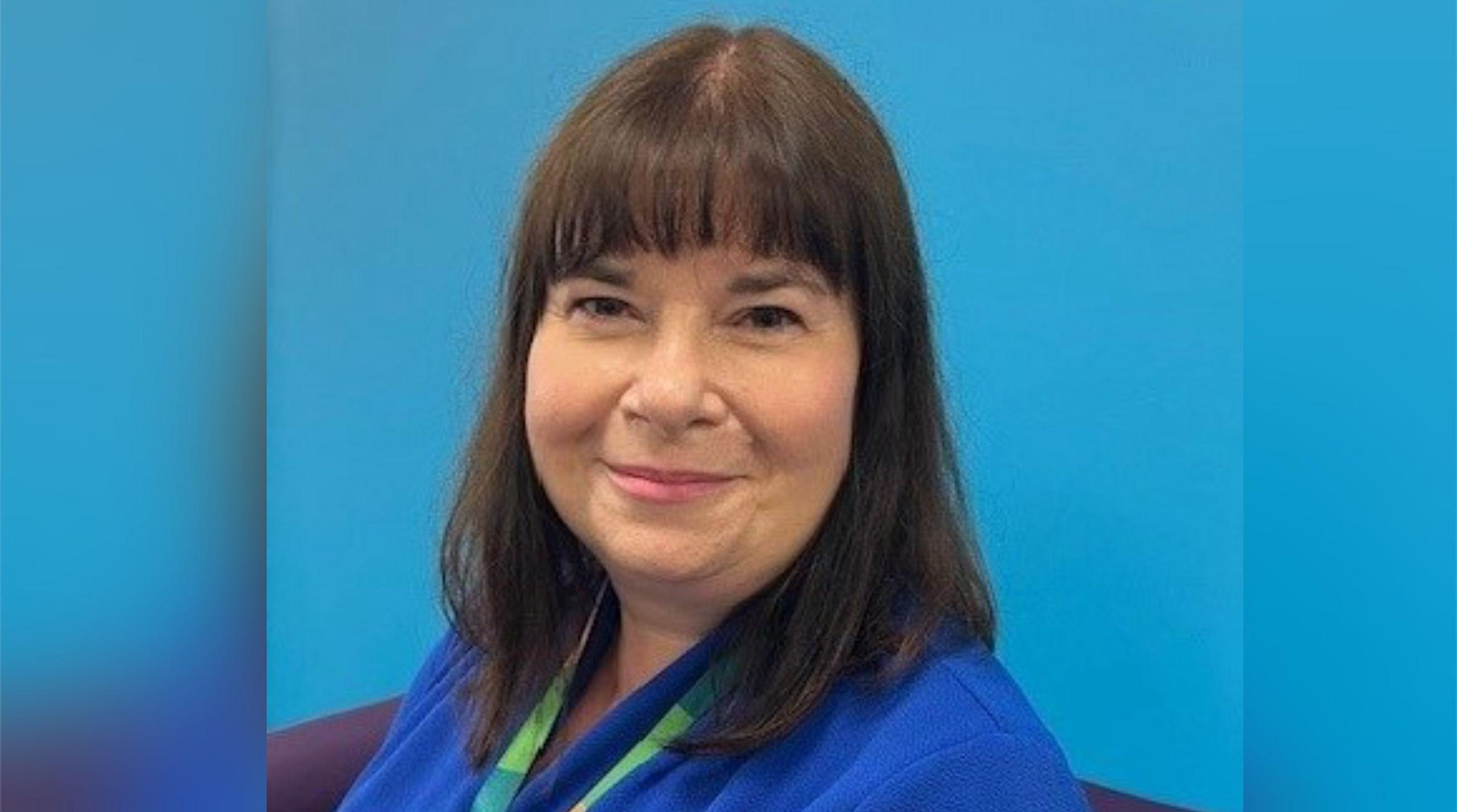 Janine Elson with long straight black hair, smiling and wearing a blue jacket with a green lanyard.  She is standing in front of a blue wall.