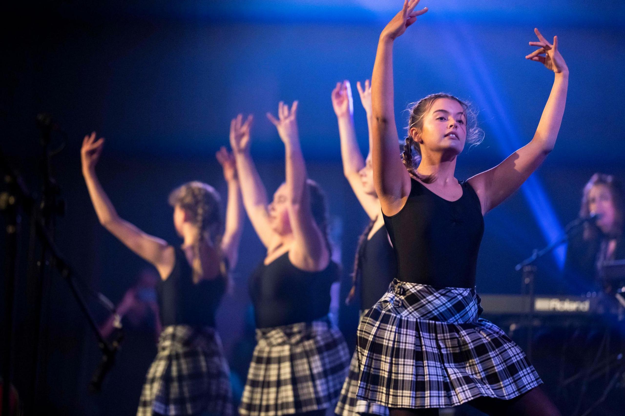 Young female dancers have their arms raised as they dance during a performance indoors. They are wearing black outfits and black and white checked tartans.