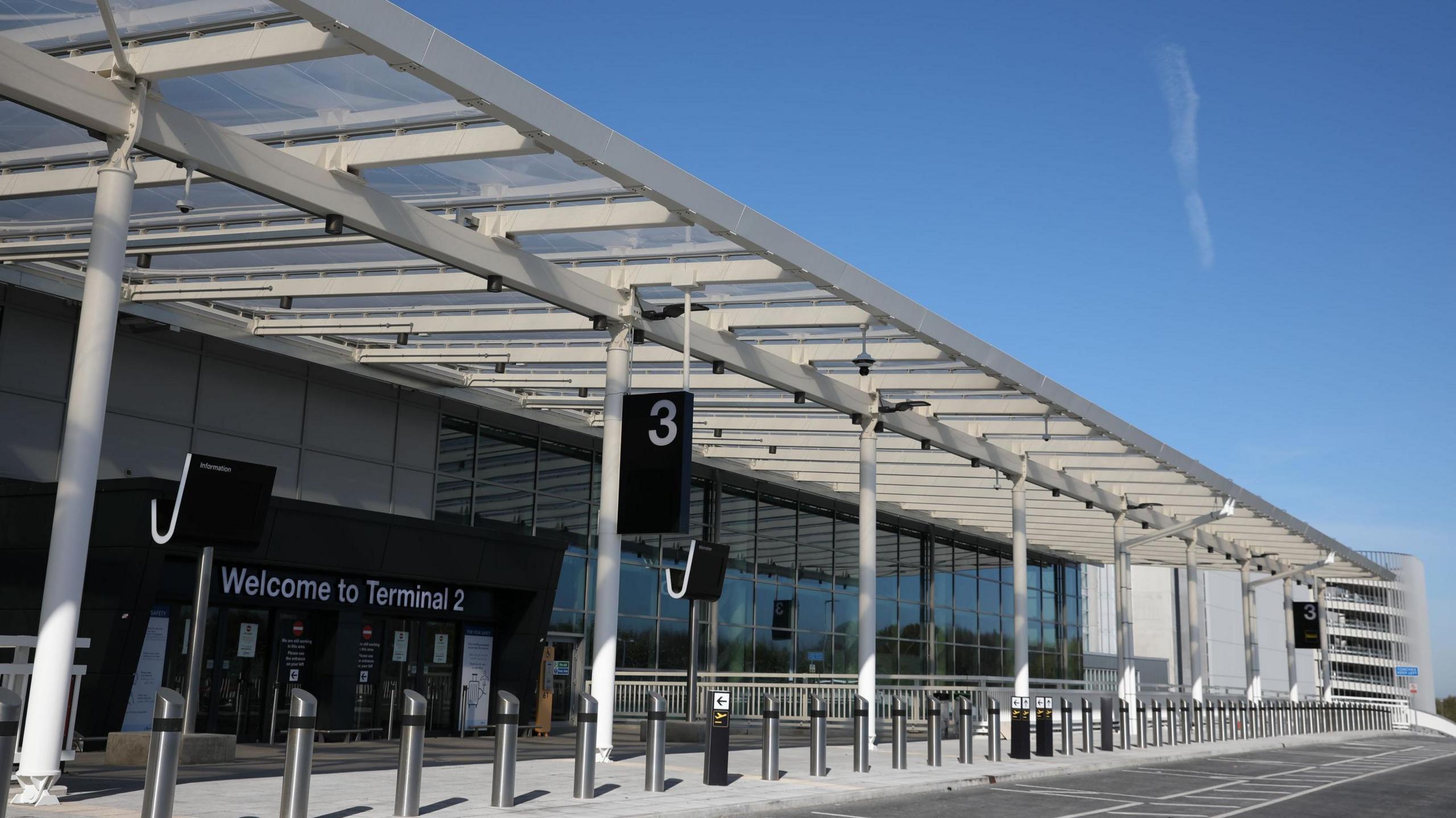 The forecourt of Terminal 2, with bollards seen running across a pedestrian area in front of parking bays. A terminal sign can be seen in front of the entrance way on a sunny day. 