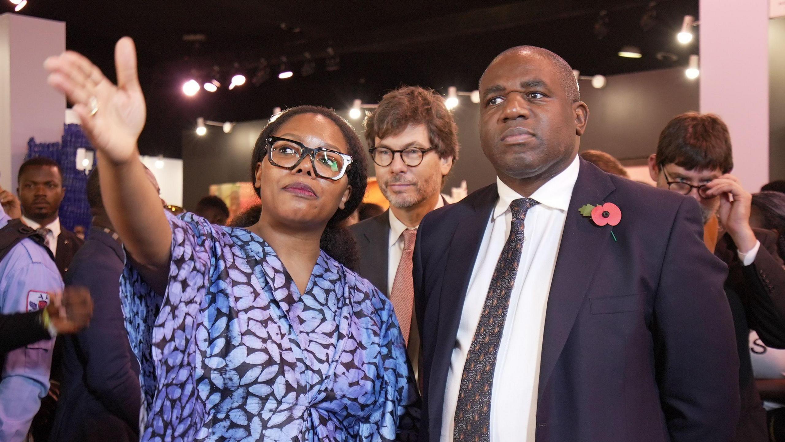 David Lammy stands beside Tokini Peterside, a woman wearing a blue dress with a leaf print and black glasses, as she points towards something off camera