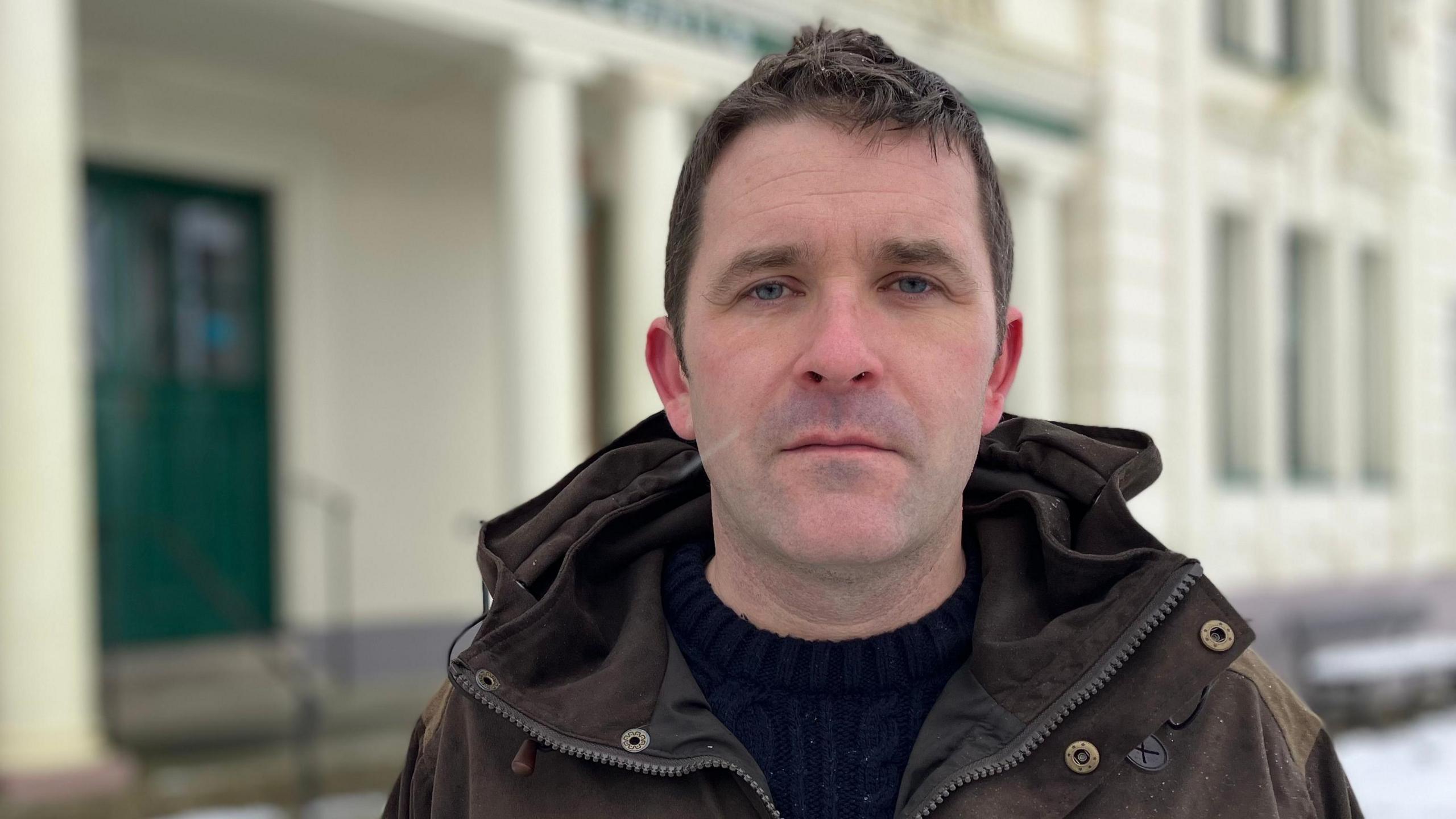 A man in a winter coat stands outside a white visitor centre building. There are columns either side of the entrance and there is snow on the ground. 