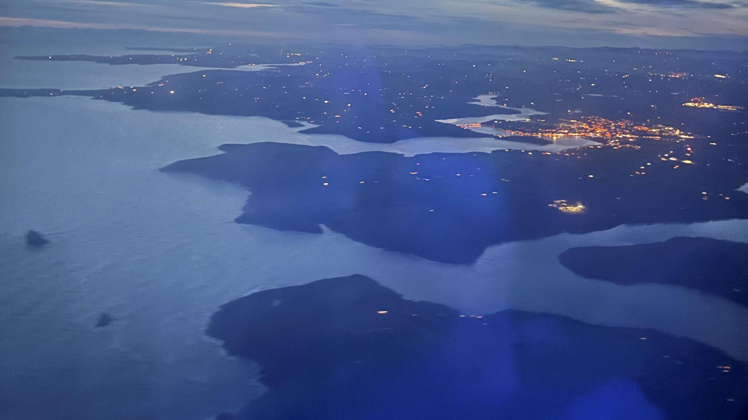 An aerial view over the east coast of Ireland taken from a plane by Andrew Wilson.  It shows a sparsely lit coastal area, a river channel and city lights in the background.