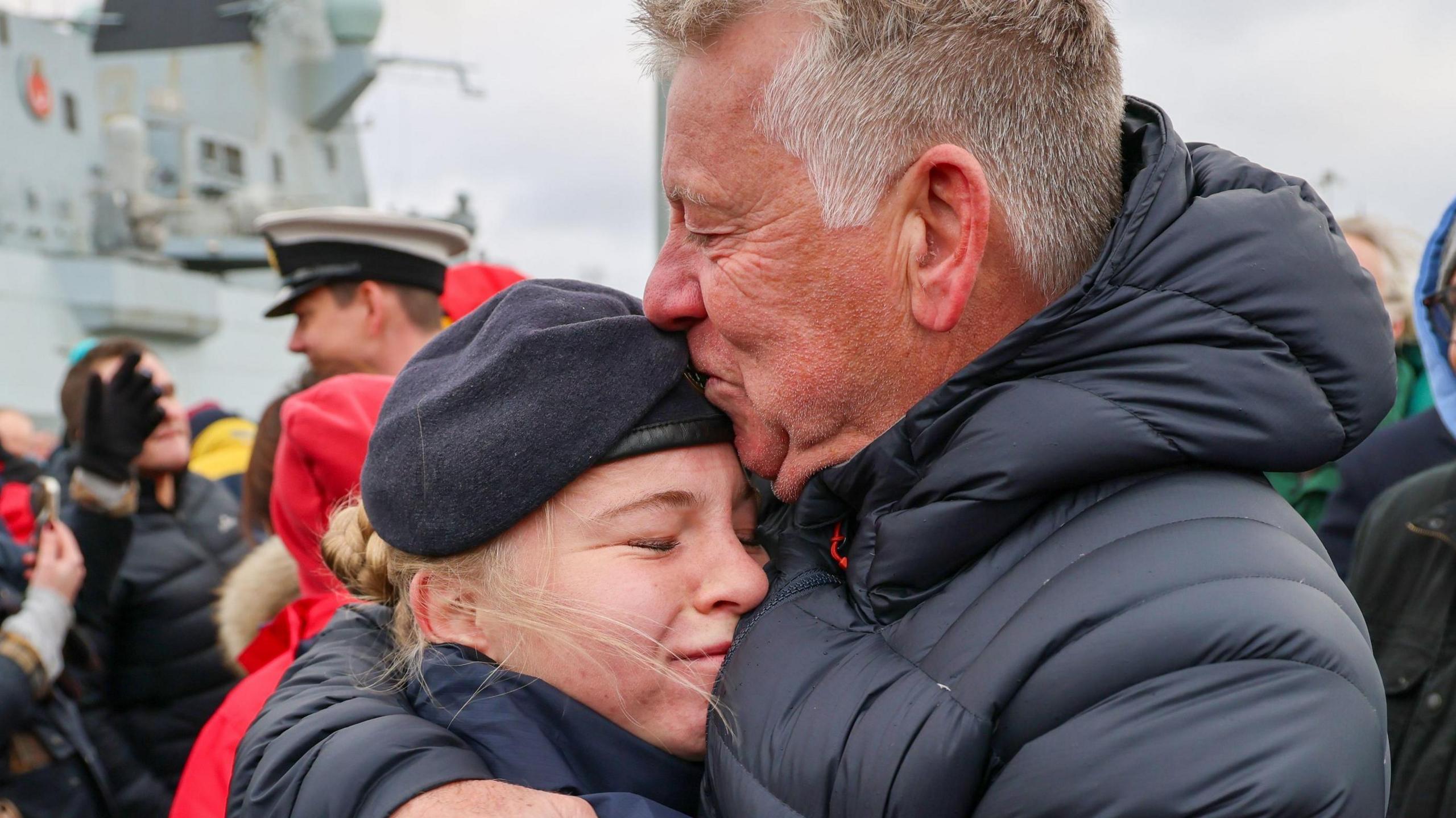 A man with blonde and grey hair, wearing a dark blue hooded puffer jacket is hugging and kissing the forehead of a young woman with blonde hair, wearing a Royal Navy beret. They both have their eyes squeezed shut and are smiling, crowds of people can be seen in the background as well as the side of HMS Duncan