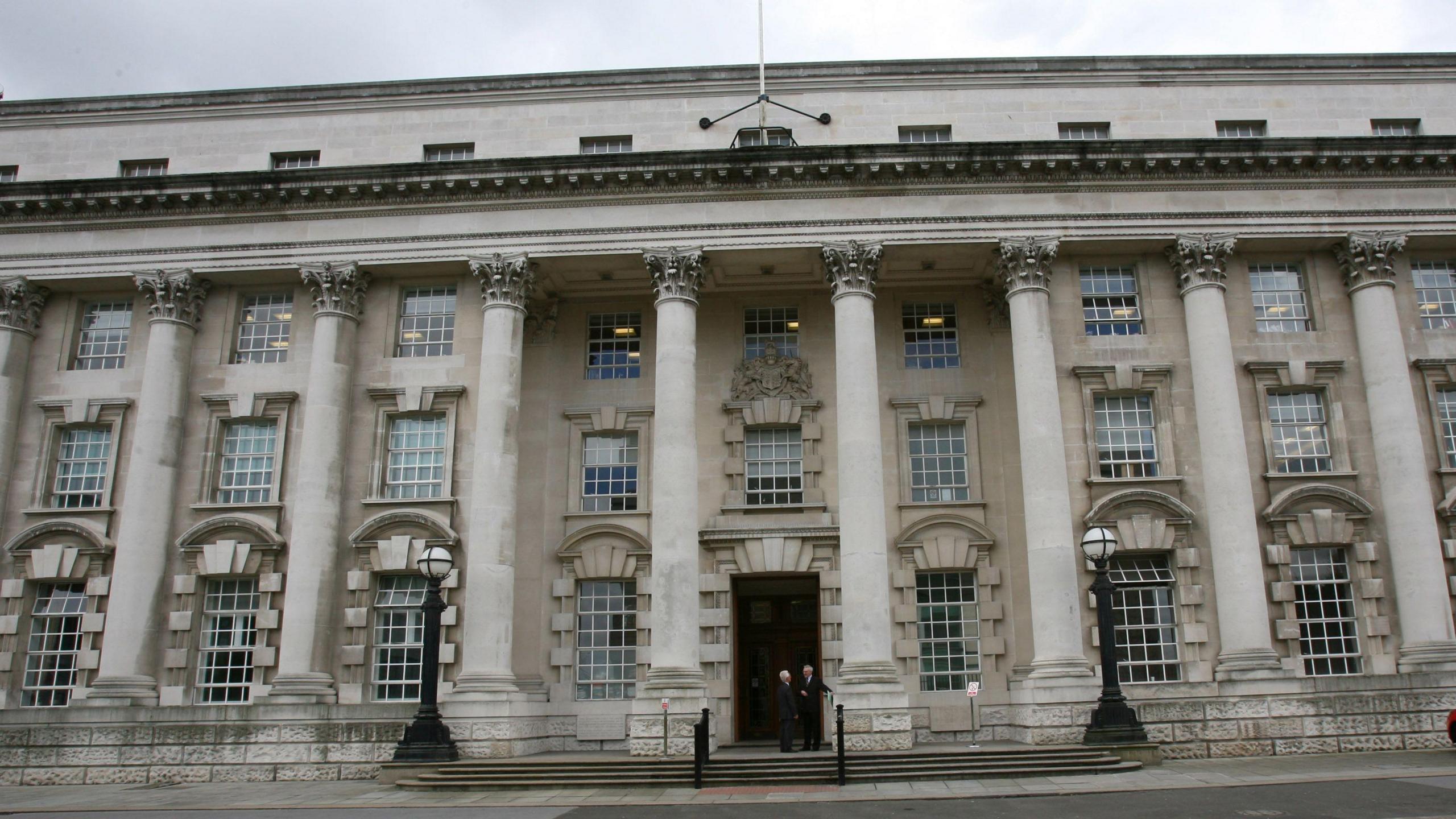 The front of Belfast Crown Court, two individuals are standing outside the front door dressed in black. A red car is parked to the right of the building. A portion of grass to the front of the building is also in view. 