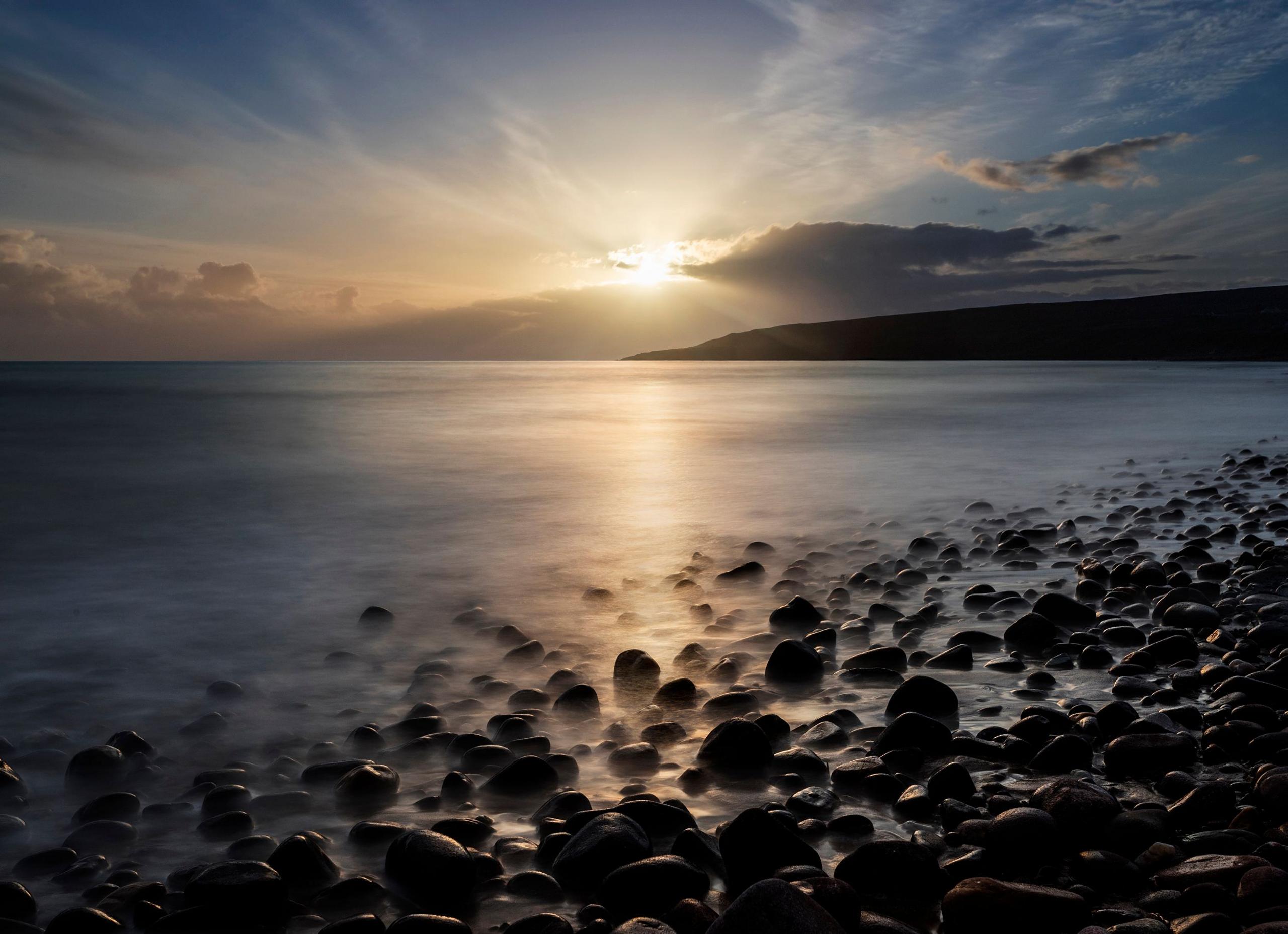 The shore of a beach with stones in the foreground and a misty background