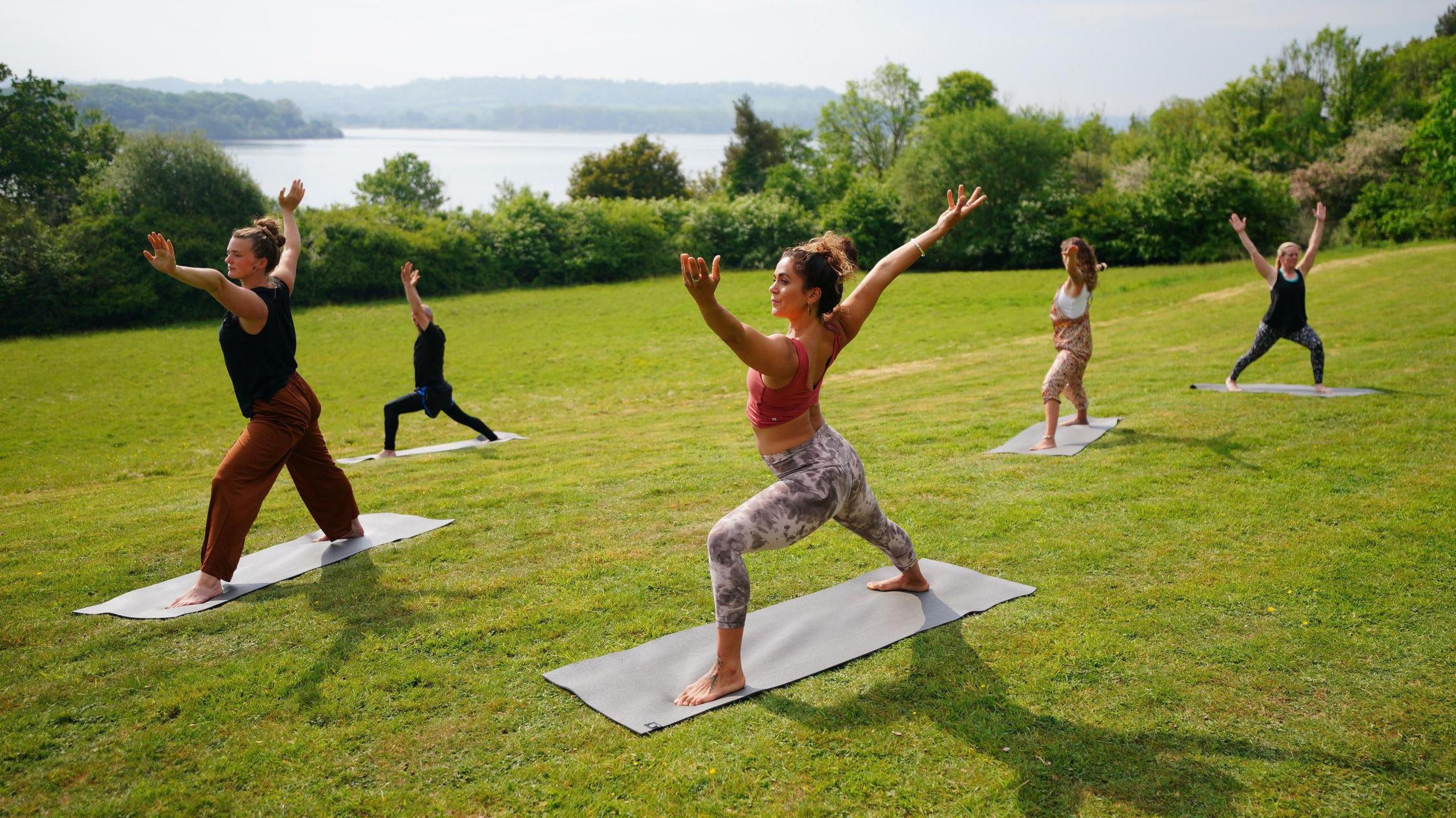 A small group of five people is spread out on a green hill with a lake in the distance. They are standing on yoga mats making yoga poses, stretching out their arms. It is sunny.