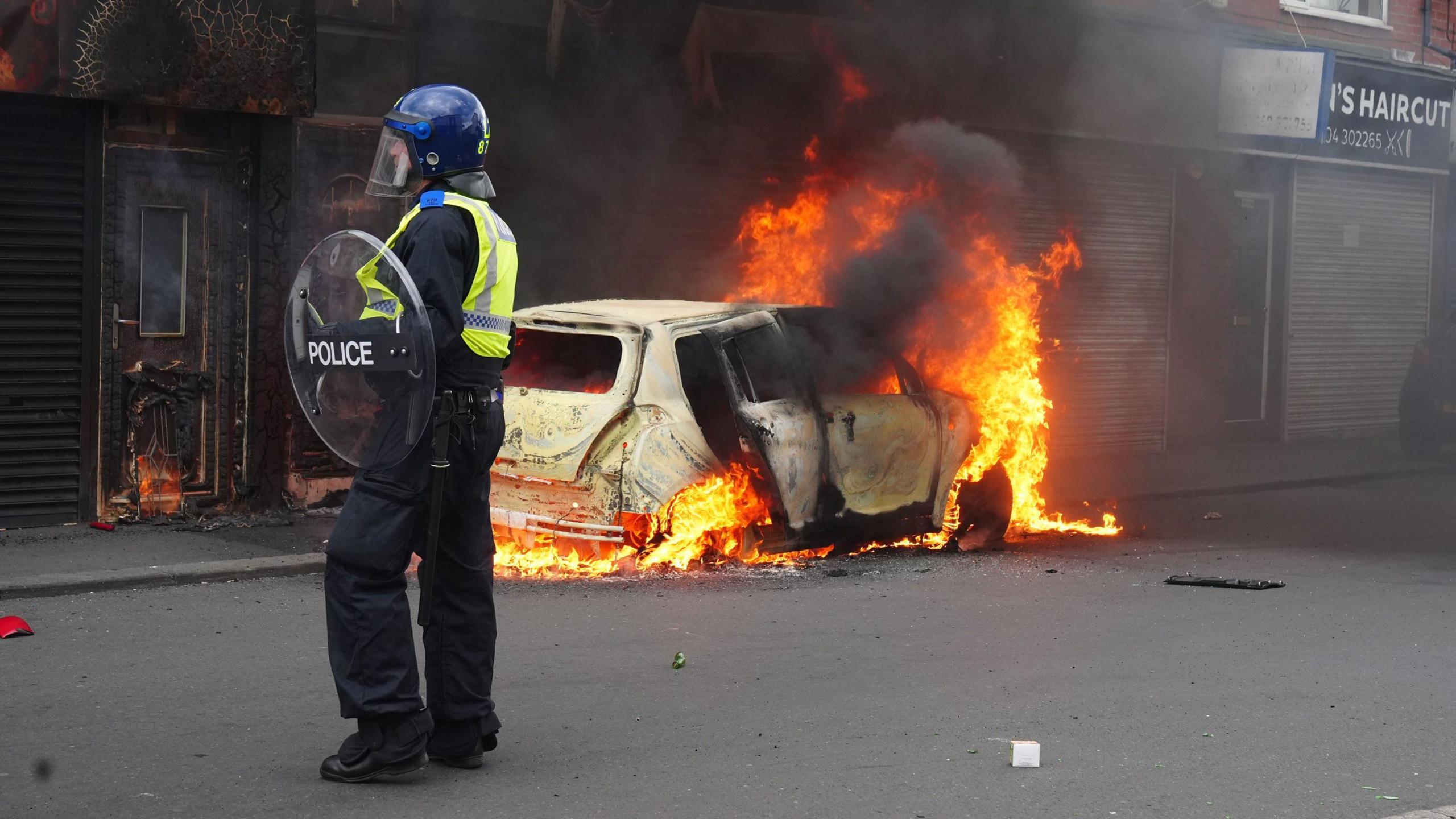 A police officer in a blue helmet and carrying a round shield stands close to a car that is totally on fire.