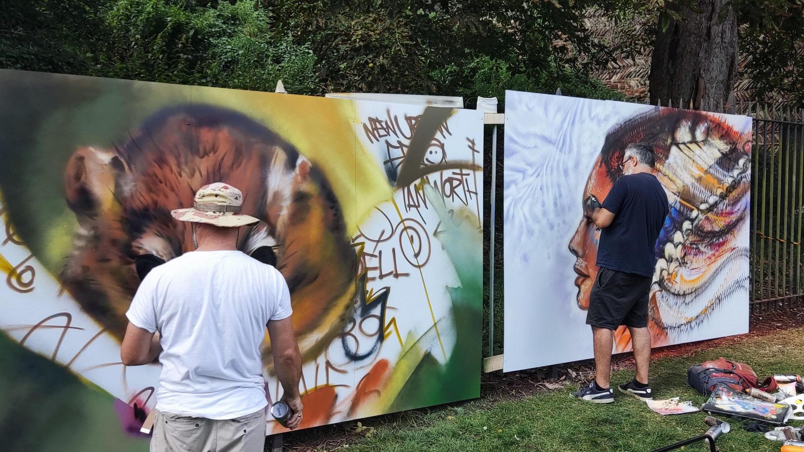 Two street artists painting murals at a previous event. Both men have their backs to the camera. The man on the left is painting a brown dormouse and the man on the right is painting a woman with feathers for her hair. Art supplies are next to his feet. 