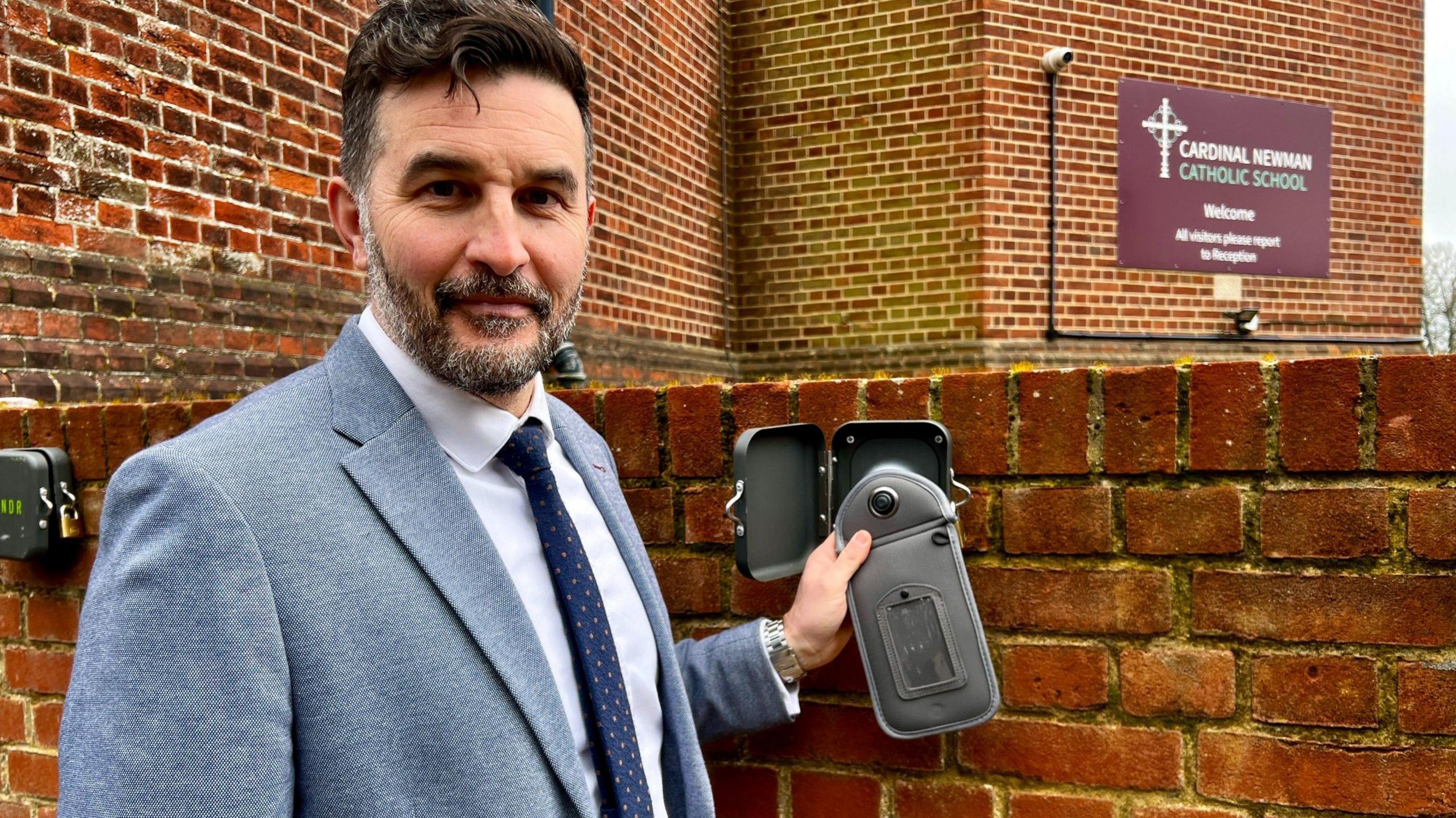 Shaun Meaney, wearing a light blue jacket, shirt and dark blue tie, holds up a pouch for a smartphone, in front of a brick wall, with the school's sign in the background. 