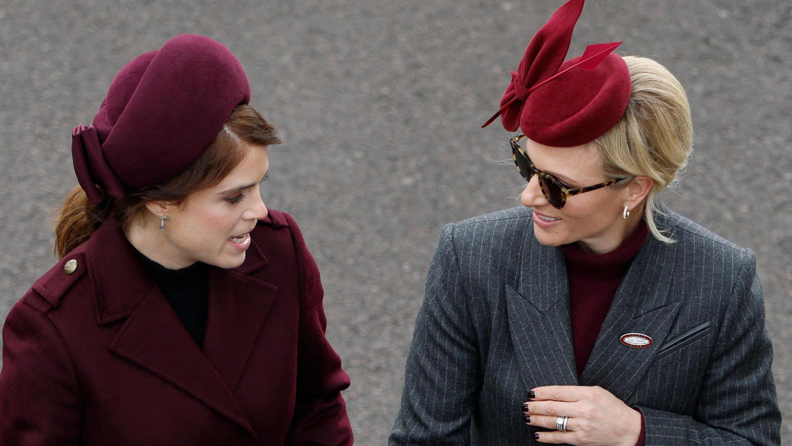 Princess Beatrice in a burgundy coat and matching hat arrives at the Cheltenham Festival course in Gloucestershire with Zara Tindall wearing a dark grey pinstripe coat and dark red hat. They're chatting to each other as they walk in to the ground. 