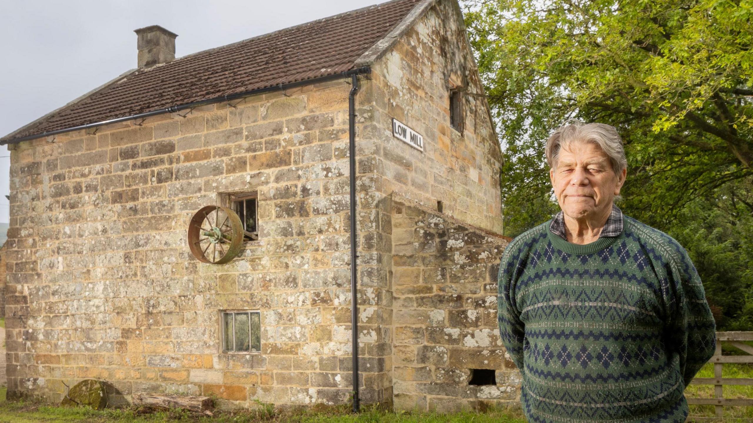 George Harrison stood outside Low Mill, an historic-looking stone building. Mr Harrison has grey hair and a green sweater and is smiling.