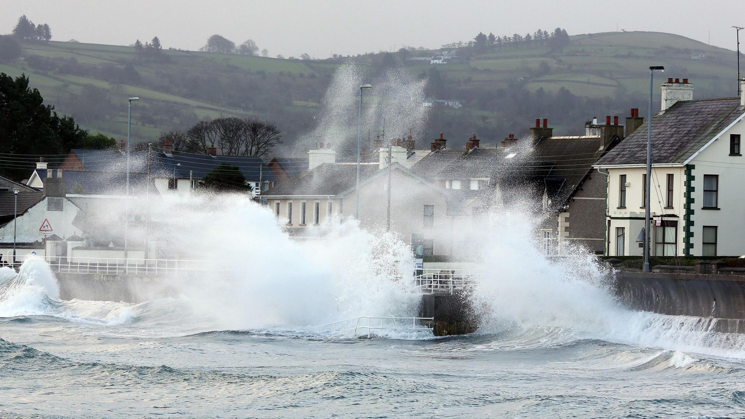 Waves crash against a promenade along the Antrim Coast in Northern Ireland - the sea is grey and buildings sit alongside the promenade. In the background green hills can be seen.