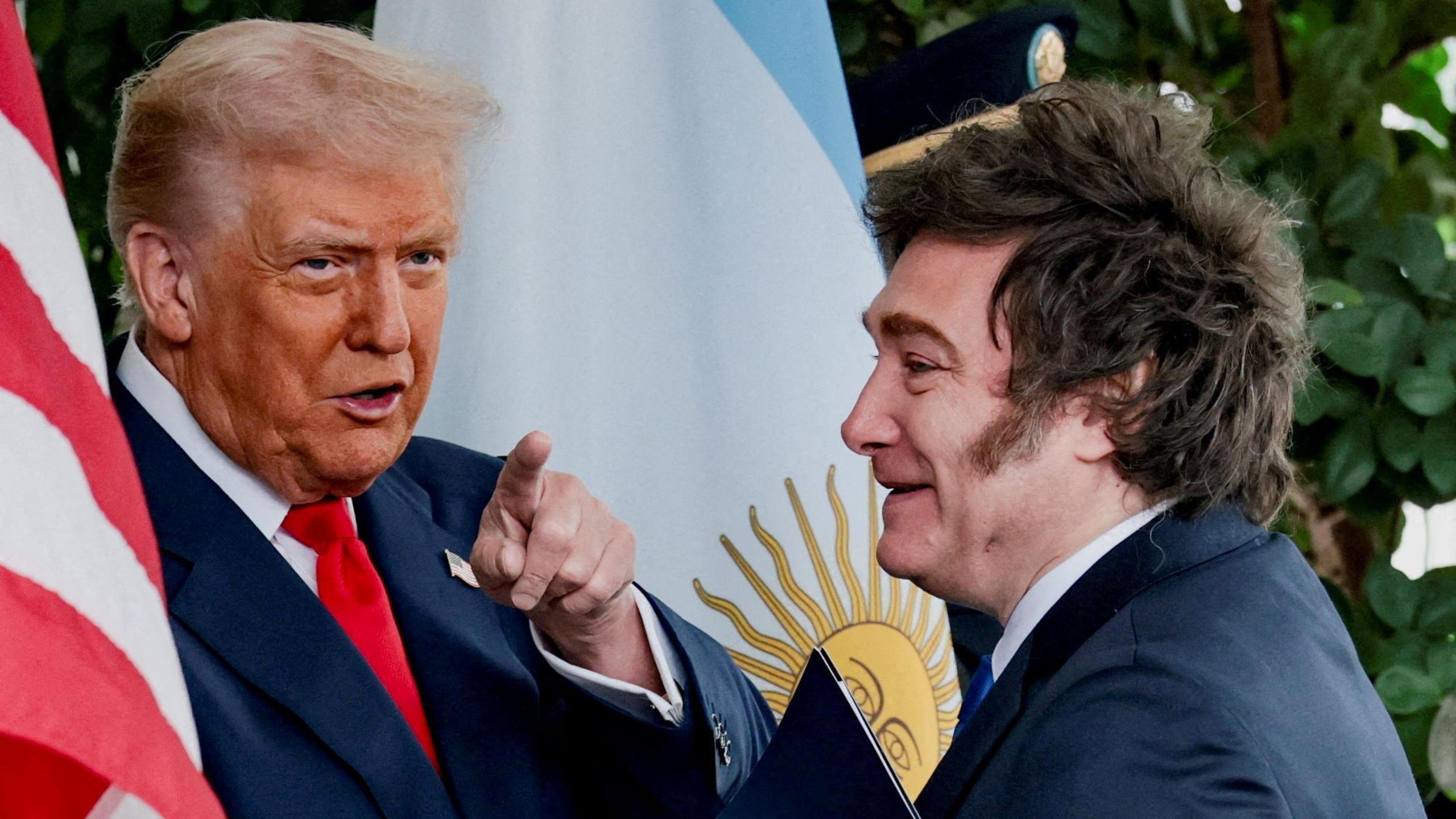 Two men wearing suits stand in front of the American and Argentinian flags. One man points toward the camera.