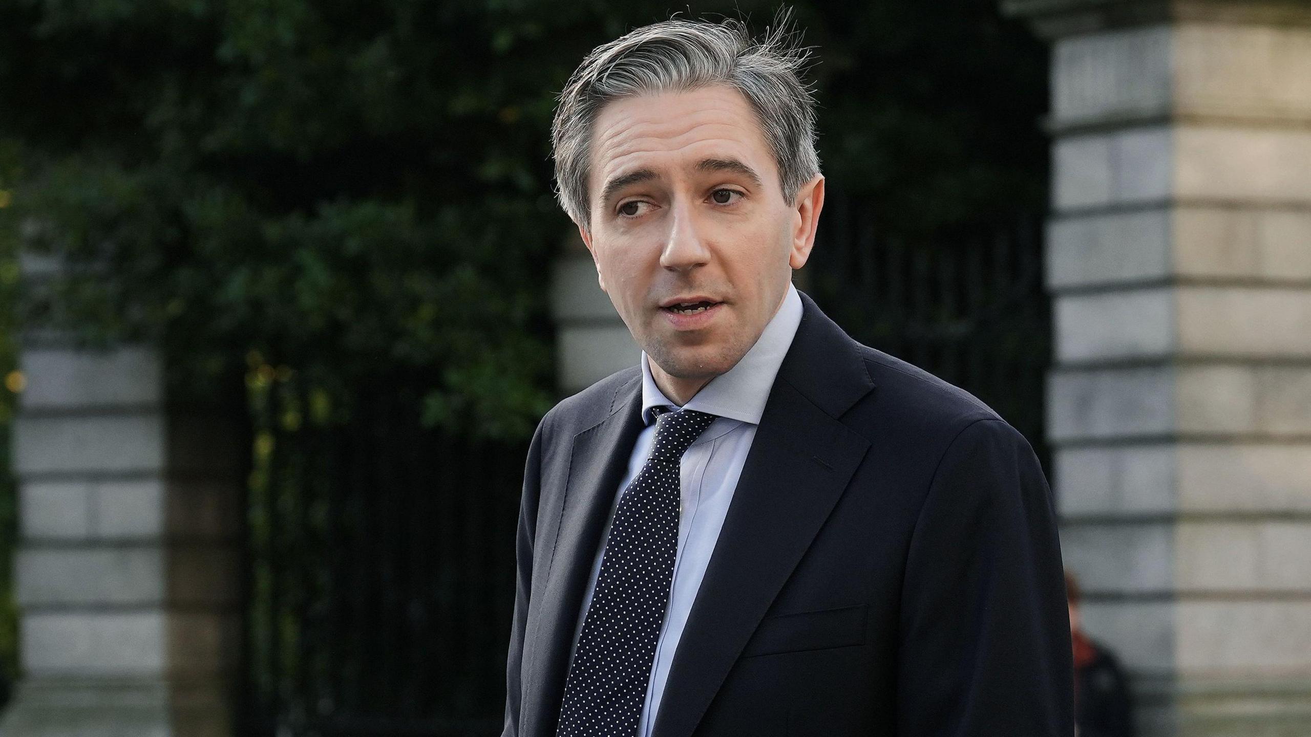 Simon Harris - a grey haired man wearing a black suit jacket, a light coloured collared shirt and dark patterned tie stands outside a light-stone wall.