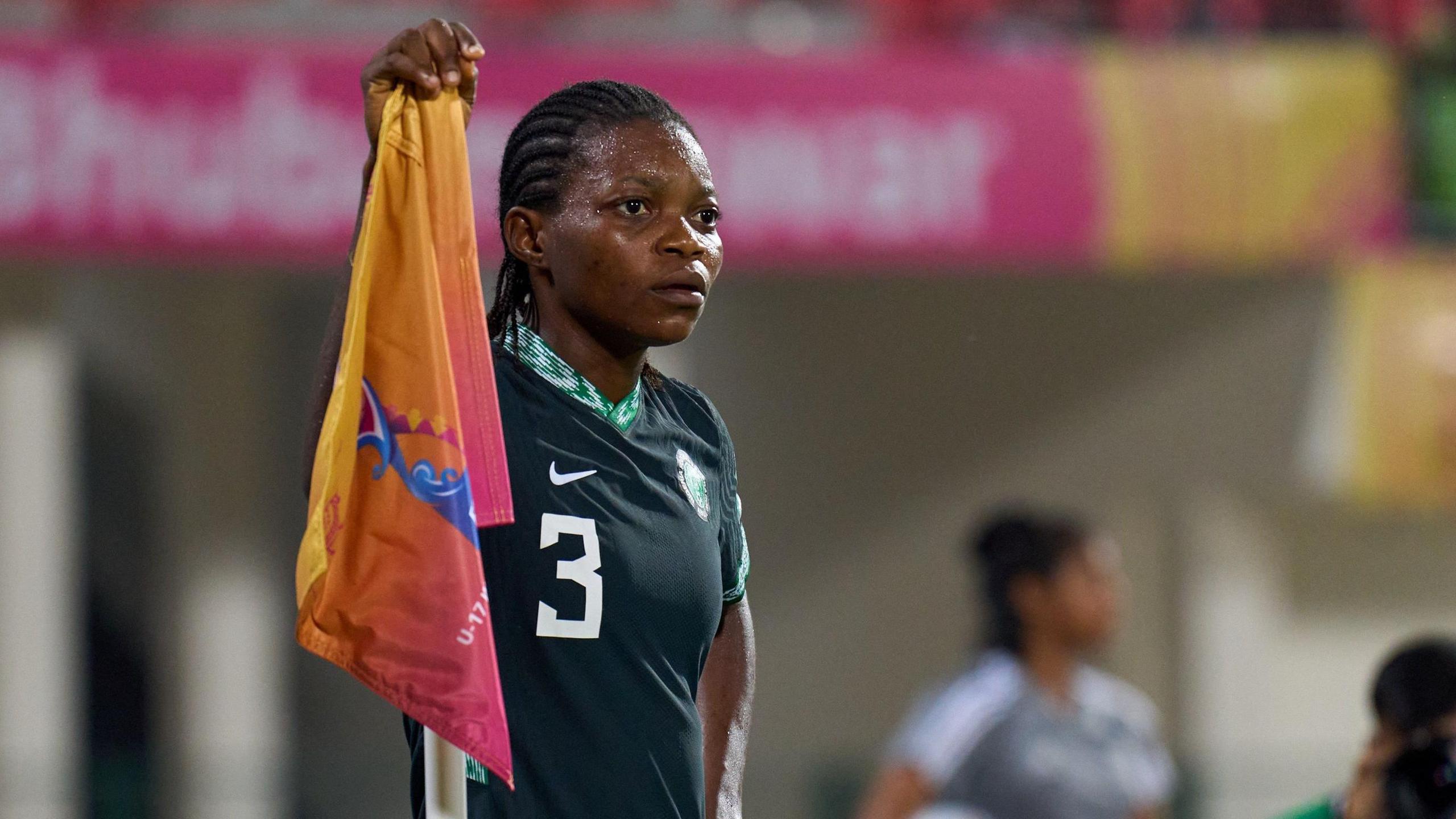 Miracle Usani, wearing a dark green Nigeria shirt with a number three on it, stands with her left hand on her hip and her right atop a colourful corner flag during a football game