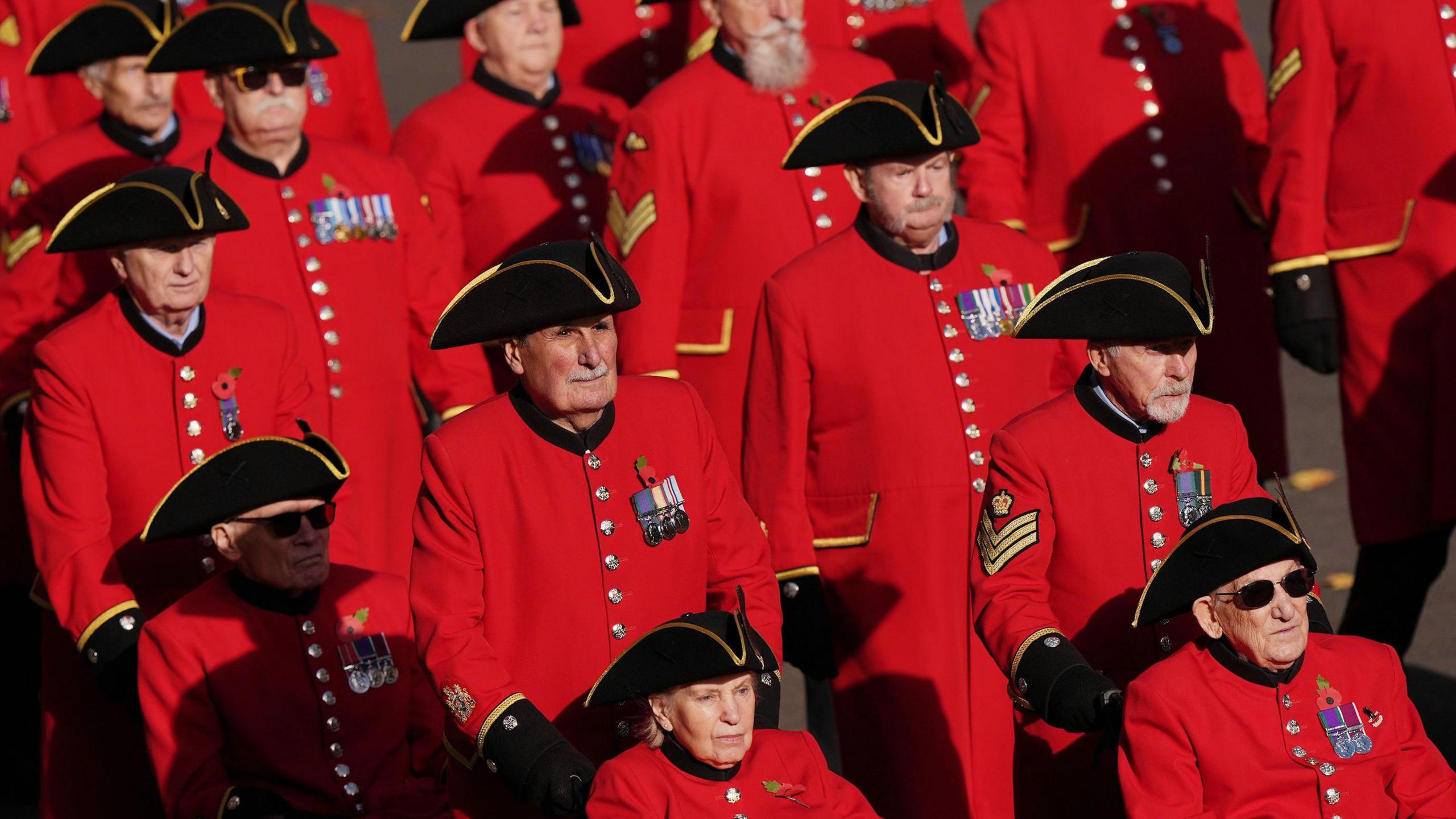 The Chelsea Pensioners in their red uniforms walking in rows. The front row shows some in wheelchairs being pushed by other pensioners.