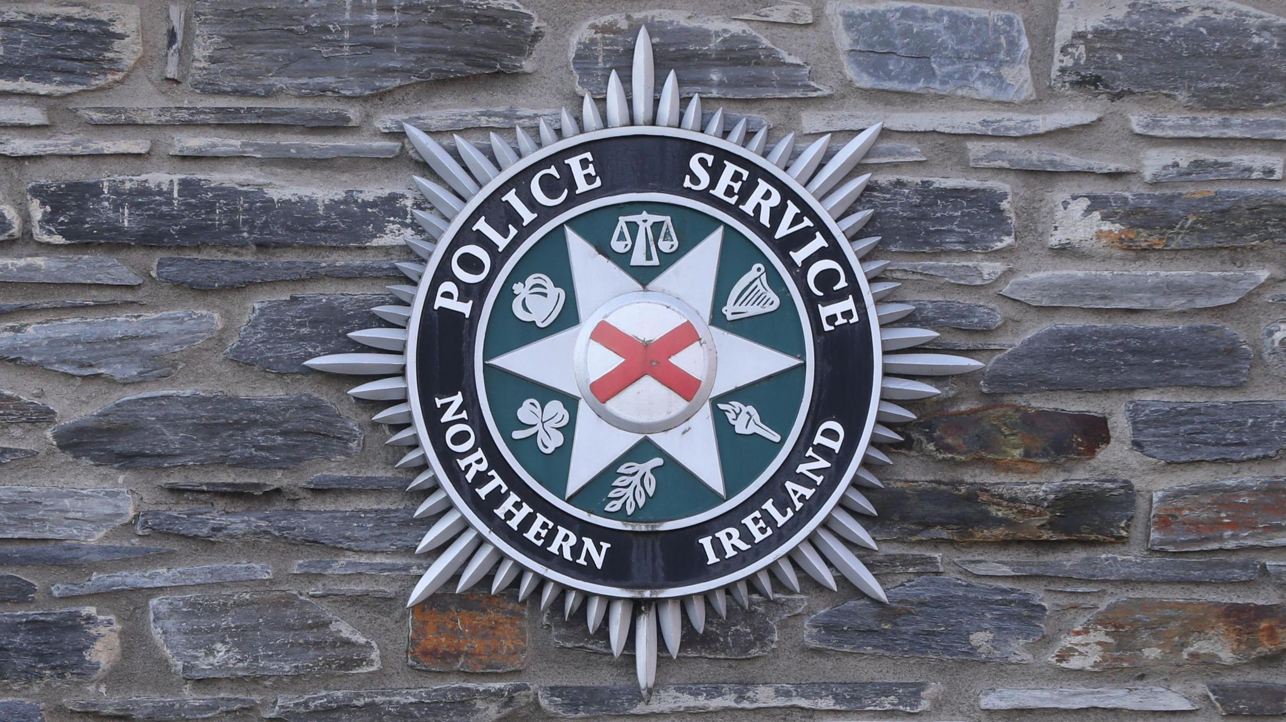 On a grey and brown brick wall is the Police Service of Northern Ireland badge. The badge is circular and it has silver spikes around it, the first section of the circle is black with white writing saying 'Police Service Northern Ireland'. The next circle inside this is green with six symbols on it in white. Then there is six white spikes with another circle inside which has a red cross.