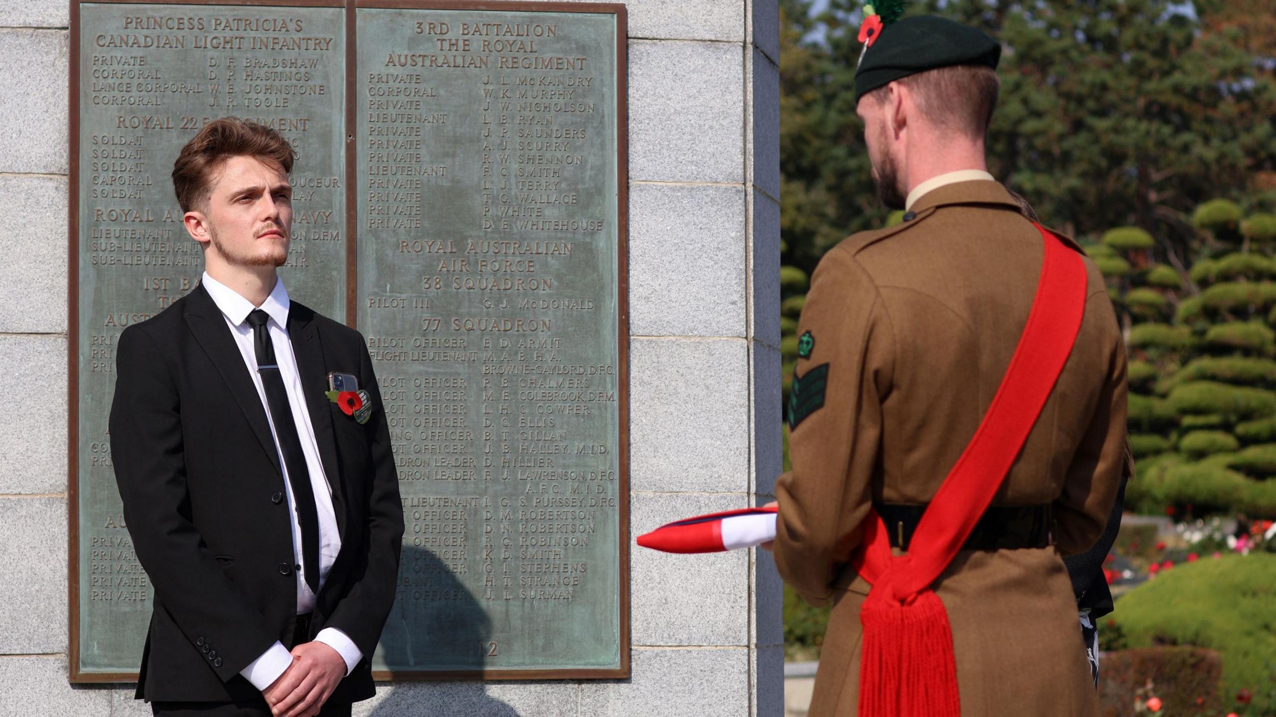 Cameron Adair at a ceremony to rename his great, great uncle's grave; a man wearing a dark suit, with a poppy in his lapel, stands in front of a memorial stone. A man in military dress, with a red sash, stands opposite him holding a folded flag.