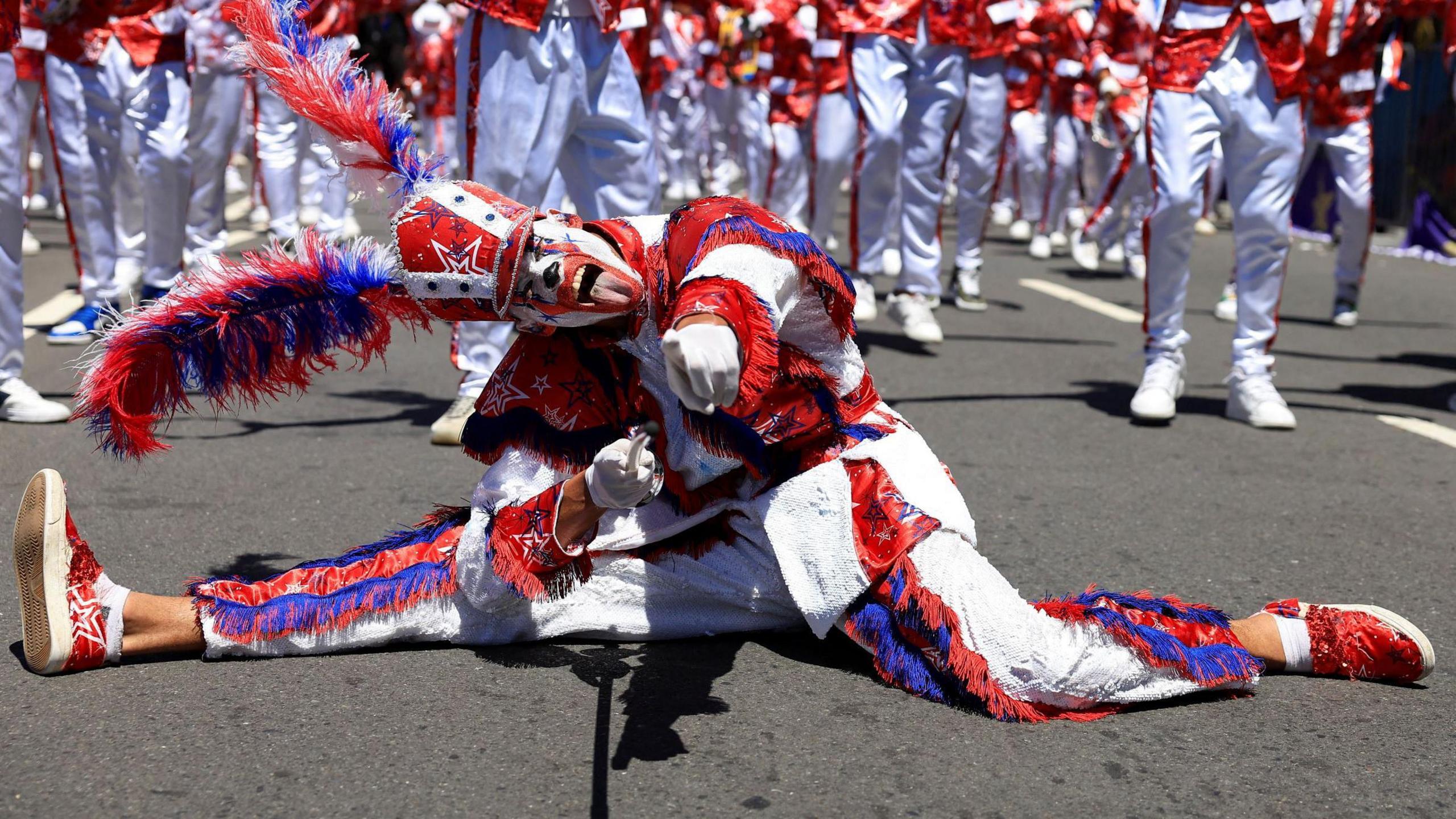 A man wearing a white, red, and blue costume with an elaborate headgear poses in a split position with his tongue sticking out at the Tweede Nuwe Jaar festival - Saturday 4 January 2025