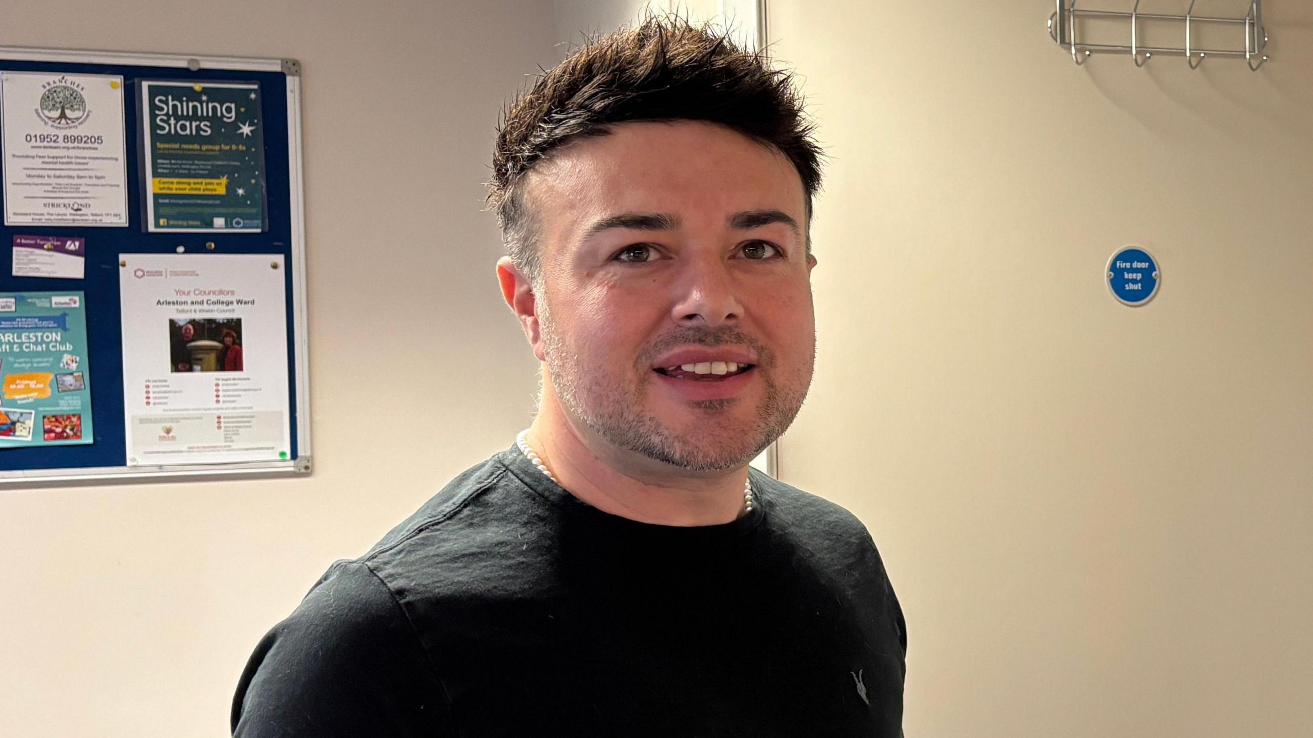 A man with short black hair and a trimmed greying beard. He is looking at the camera in a room at a community centre.