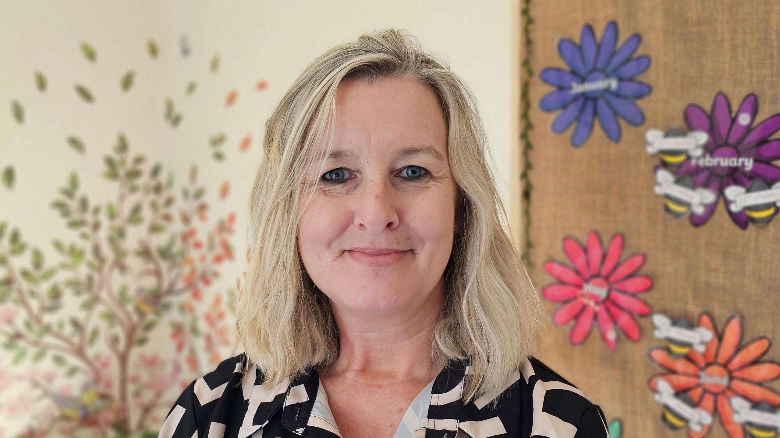 Lisa Spinks in her nursery, standing in front of a wall decorated with painted flowers. She has blonde hair and is wearing a black and cream patterned top.