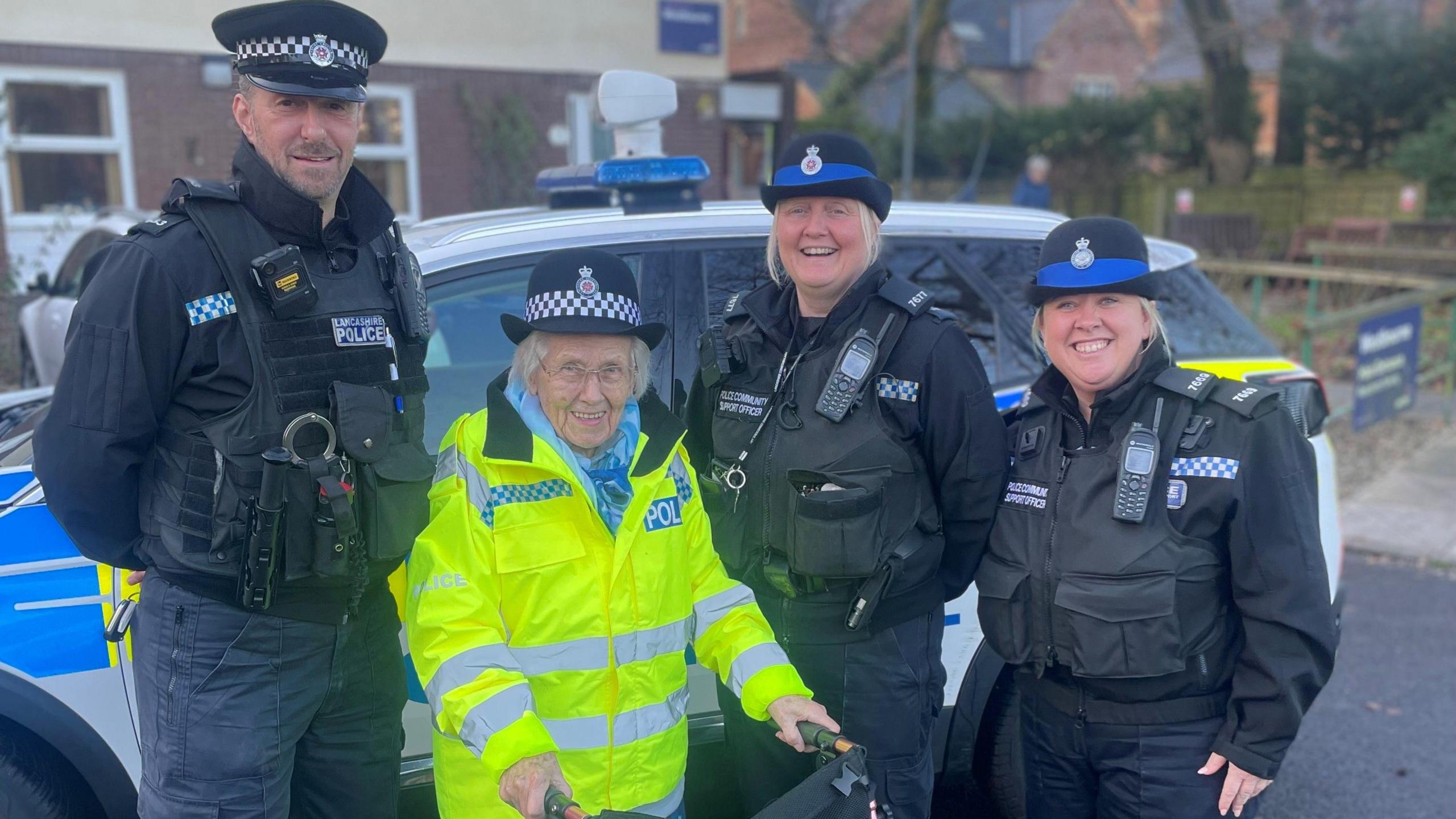 An elderly lady wears a hi-vis police jacket and police hat and smiles at the camera, as she stands with a frame in front of a police car, along with three other police officers