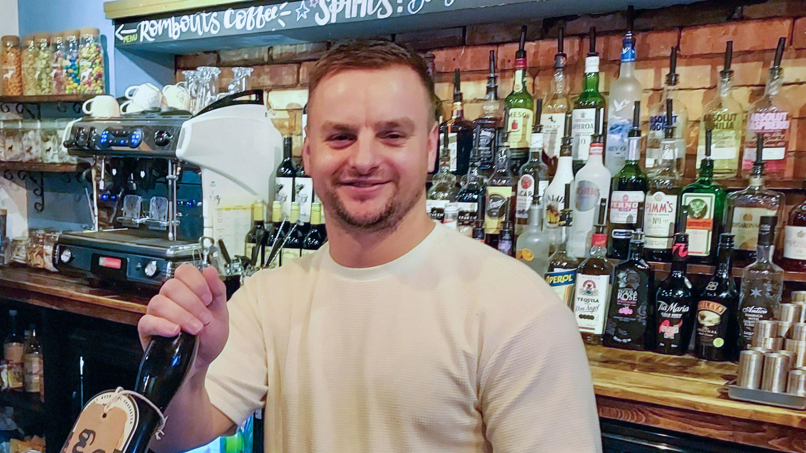 Rob Hardie with brown hair and white t-shirt pulling a pint behind the bar with alcoholic drinks and a coffee machine in the background.