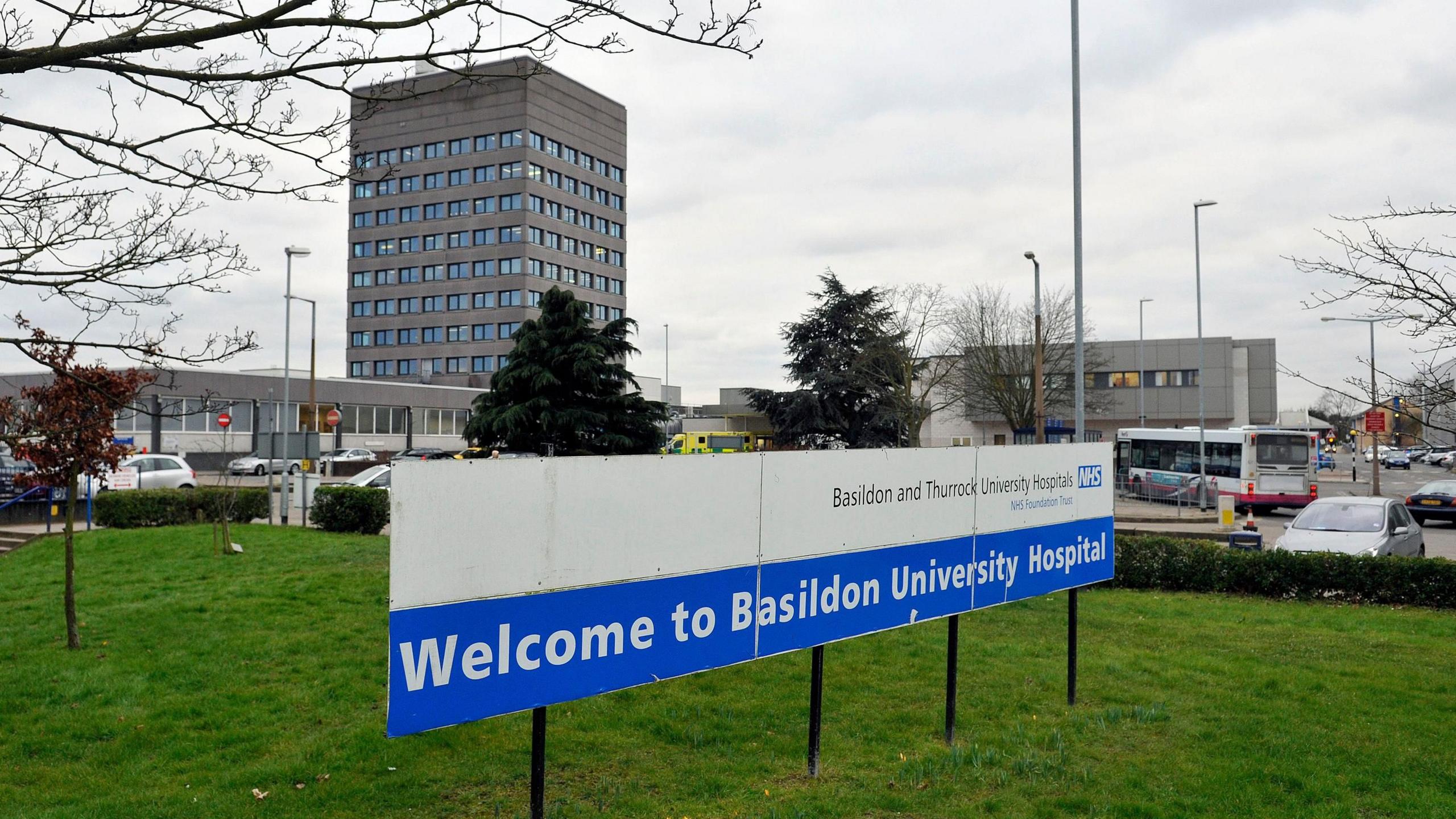 A white and blue sign that says "Welcome to Basildon University Hospital" and is placed on grass in front of the hospital car park, which has cars and a bus moving through it. In the background is a large, grey tower block with many windows.