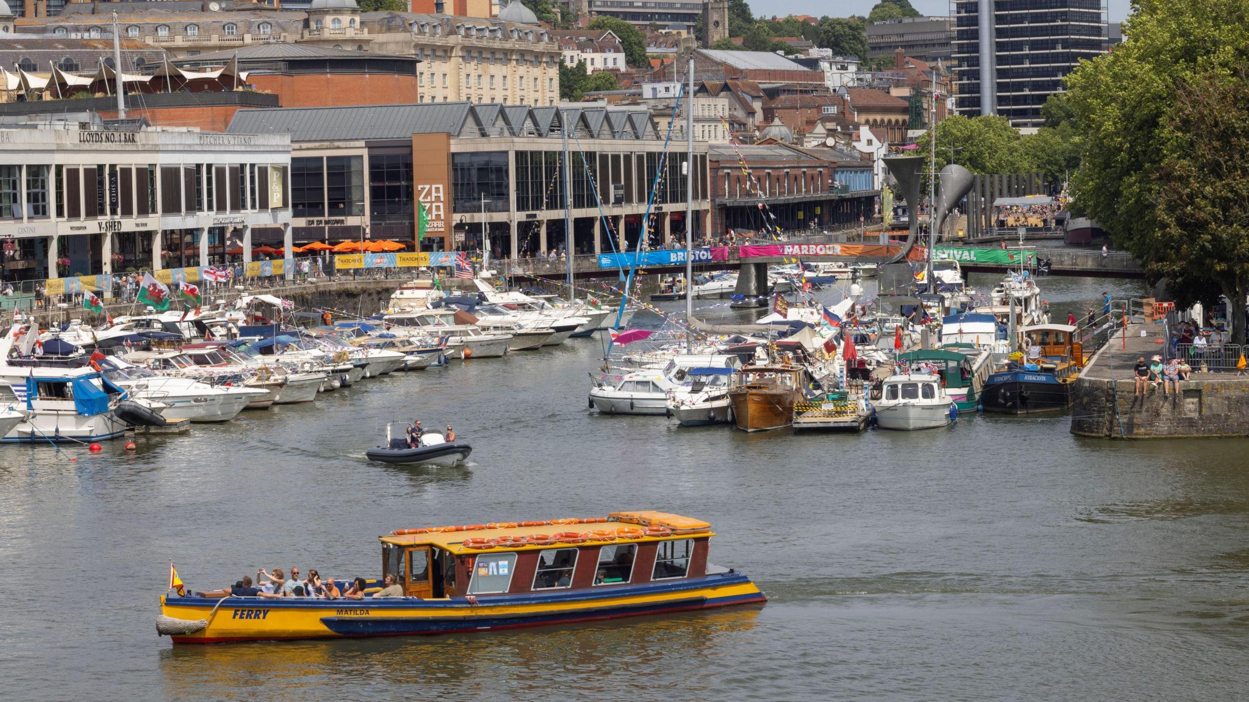 A yellow Bristol Ferry boat in the foreground on the harbour with many other boats and buildings behind it