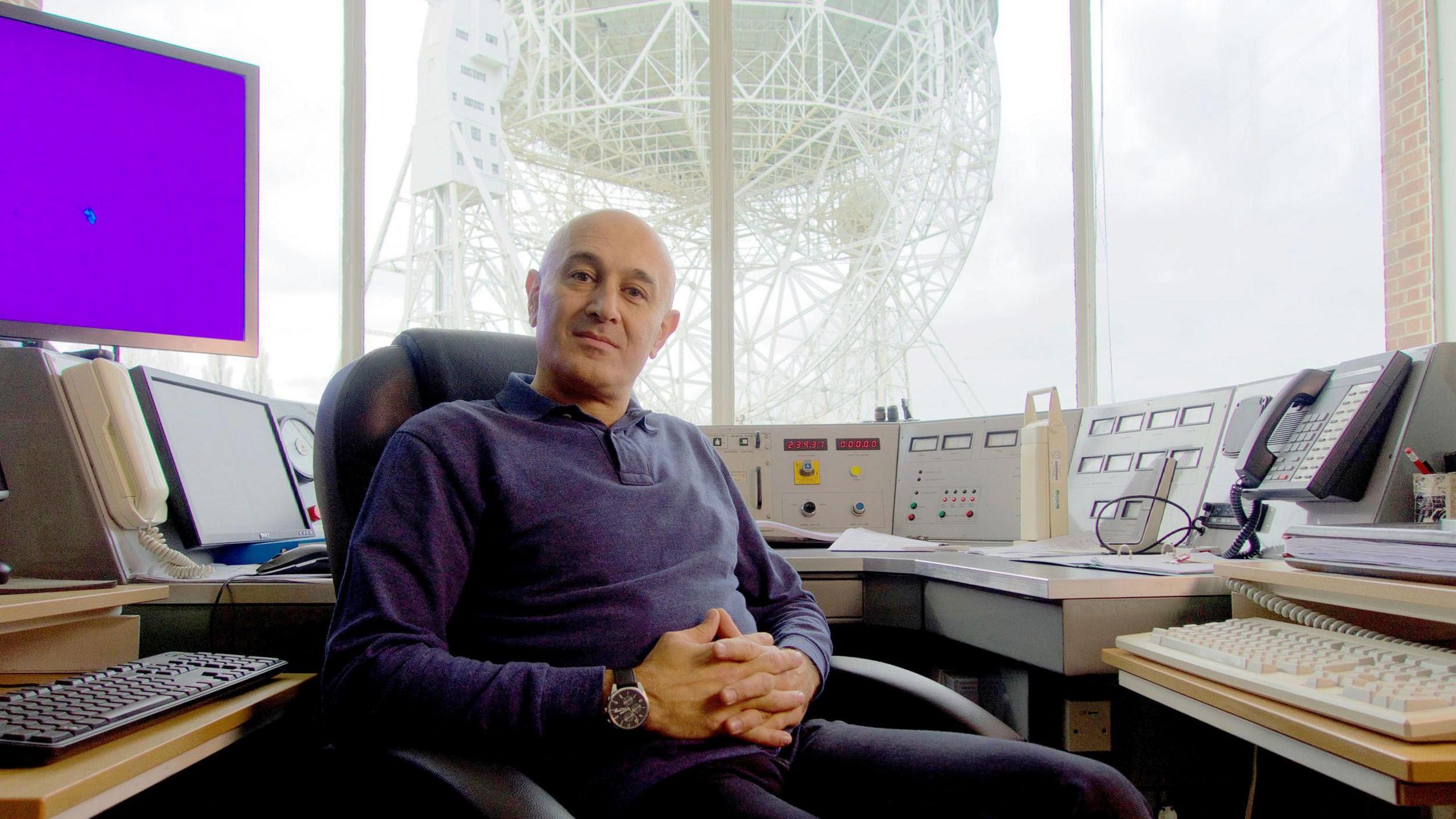 Prof Jim Al-Khalili, wearing a blue sweater and black trousers, sits hands crossed and facign the camera at the control desk of the Lovell Telescope, which can be seen out a window behind him