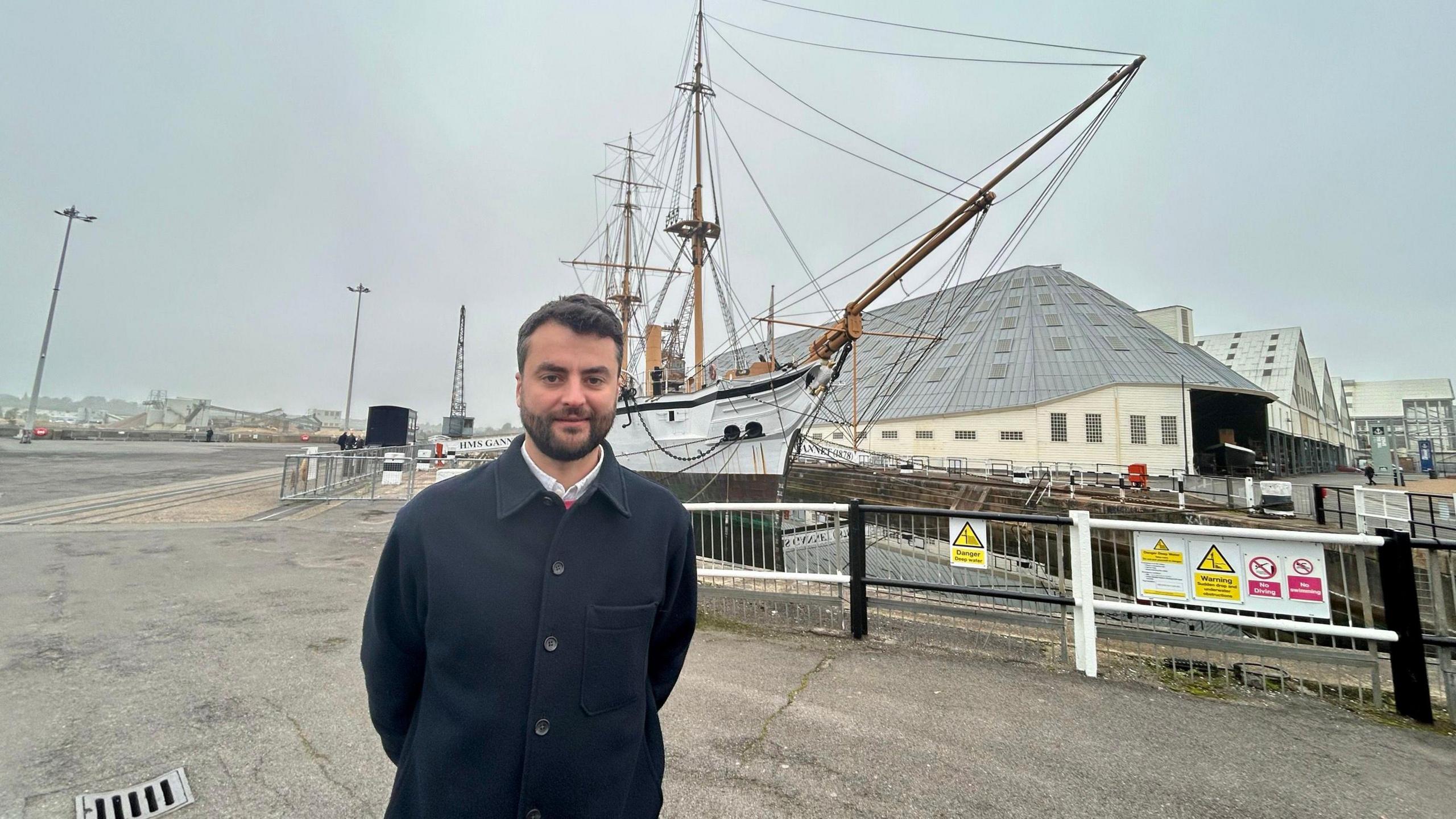 Nick Ball, collections, galleries and interpretation manager at the Historic Dockyard, Chatham, wearing a blue overcoat and a white shirt, standing in front of a sailing ship at the dock yard