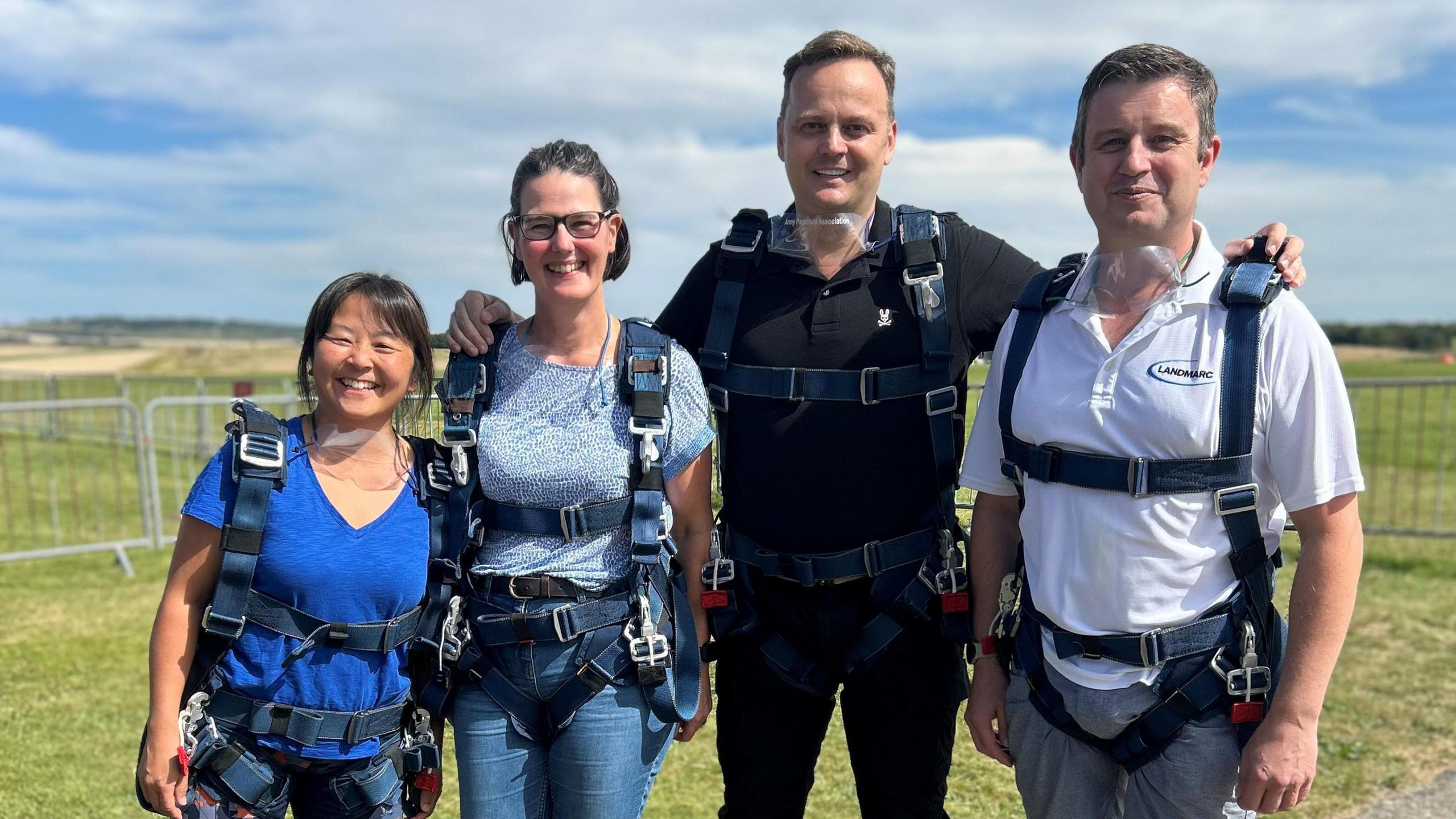 Matt (far right) stands in a field with three colleagues wearing skydiving harnesses.