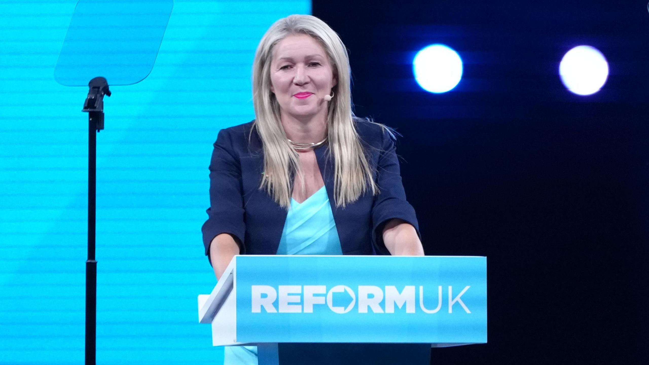 Laura Anne Jones stood in front of a podium with the Reform UK logo, in the party's teal colour. She has a dark blue blazer and a light blue dress on.