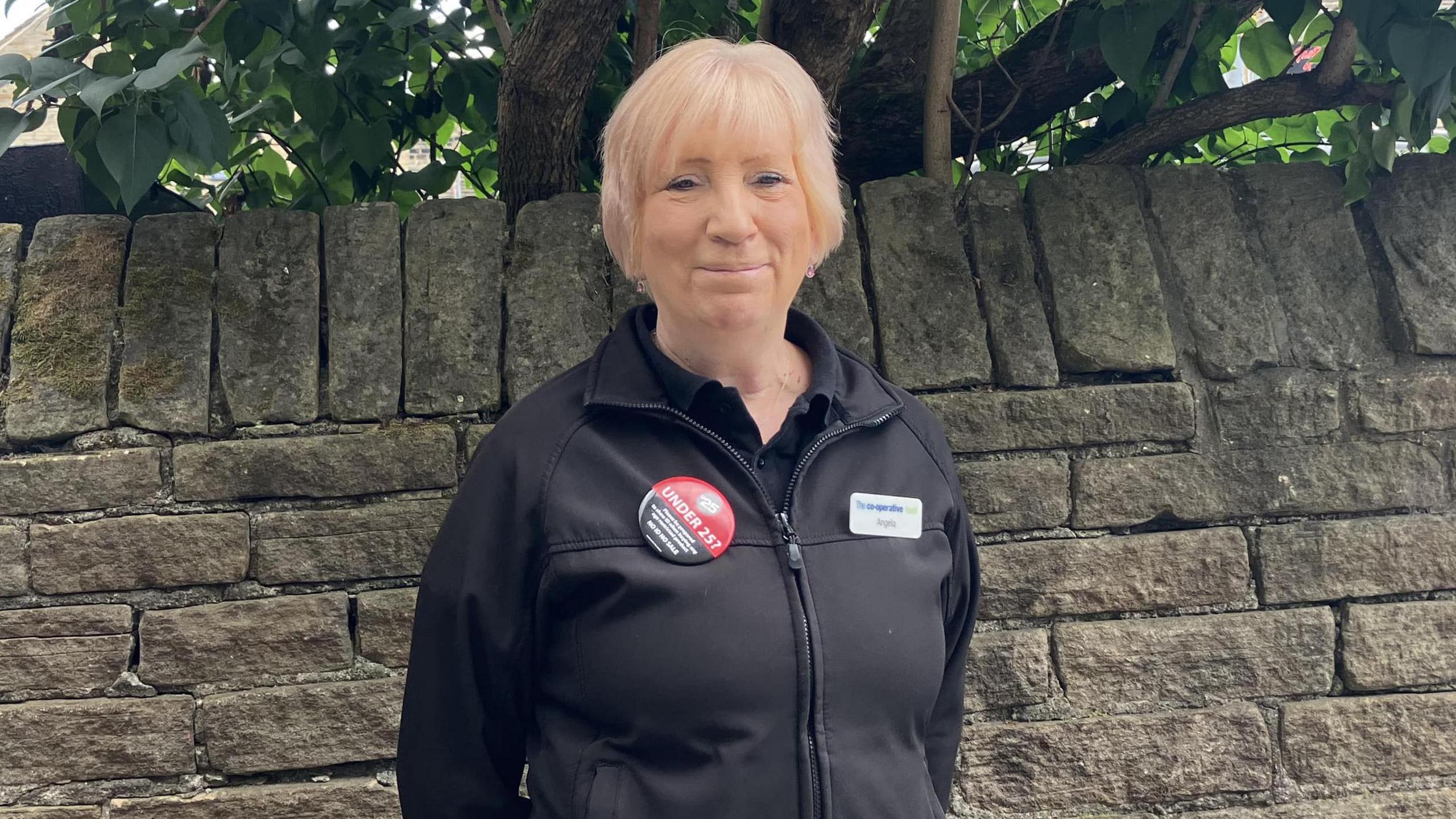 A woman in her sixties, with fair hair. She is wearing a black jacket with a couple of badges on. Behind her is a stone wall and a tree.